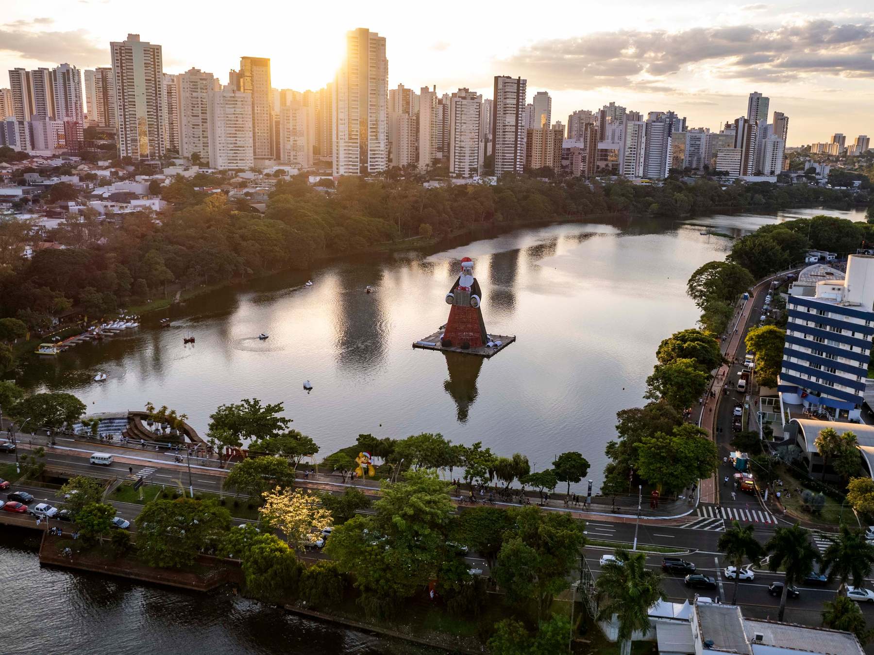 Vista aérea do Lago Igapó ao pôr do sol, com os prédios da Gleba Palhano contra o céu dourado, a água refletindo a luz e uma decoração de Natal flutuando no meio do lago, em Londrina.