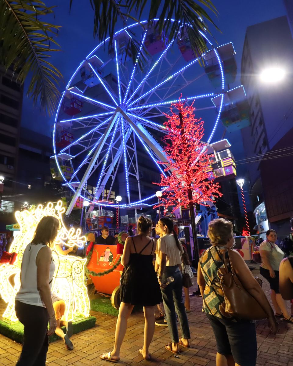 Roda-gigante iluminada de azul e árvore de luzes vermelhas no Calçadão de Londrina, com pessoas passeando à noite.