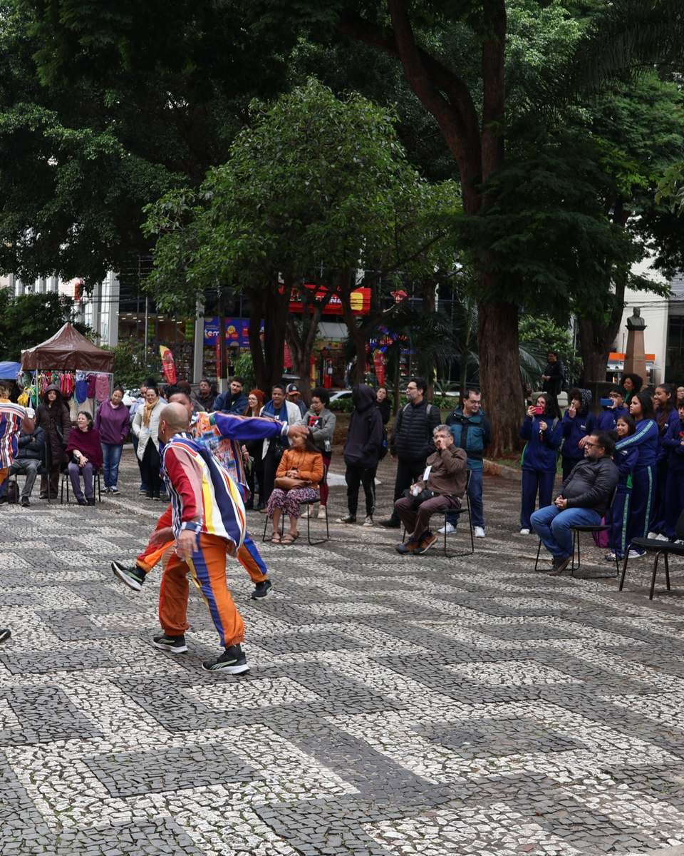 Artistas de roupas listradas coloridas se apresentando no Calçadão de Londrina durante o FILO, com o público assistindo sentado e em pé.