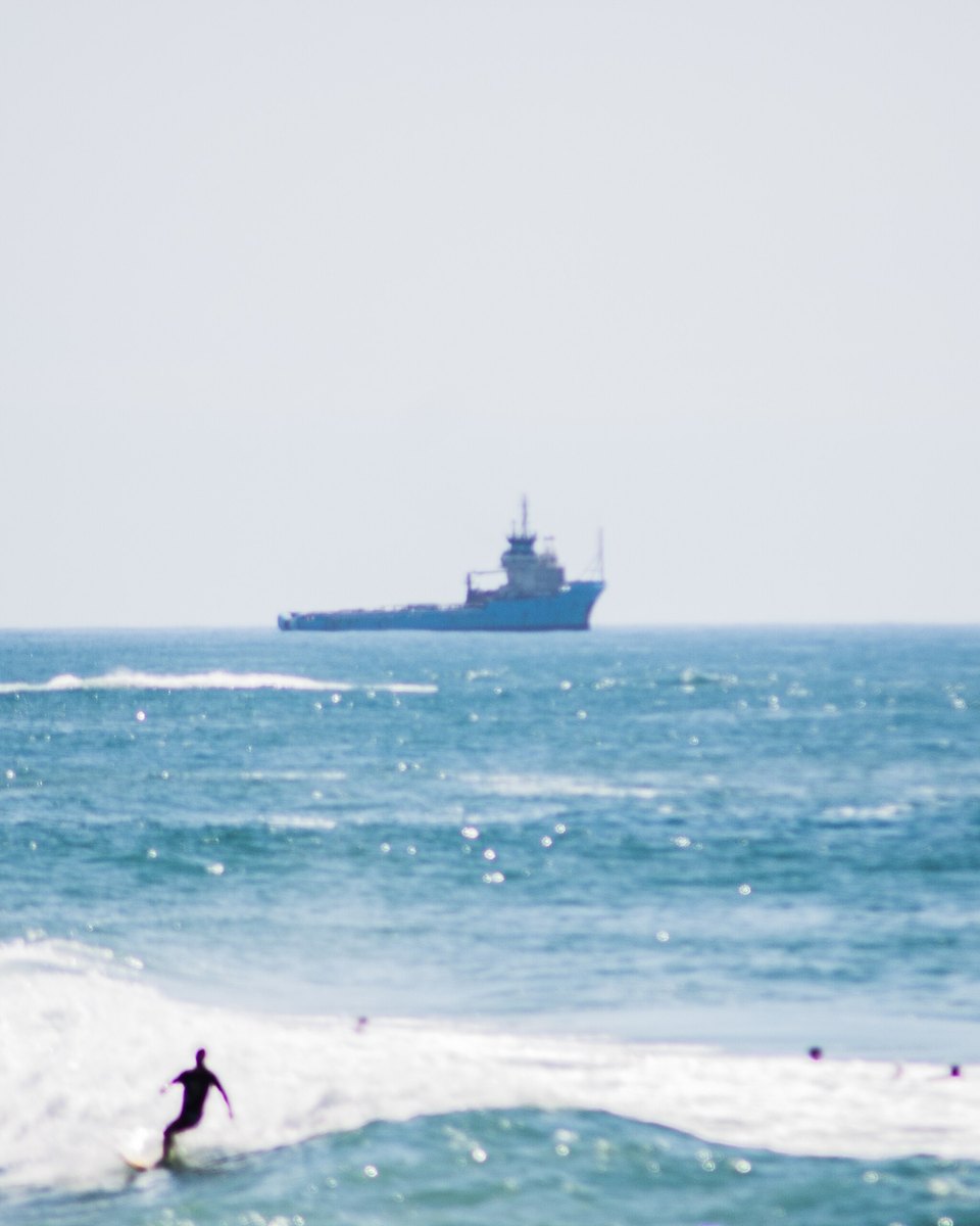 Surfista numa onda em Macaé com um barco de apoio offshore no horizonte.