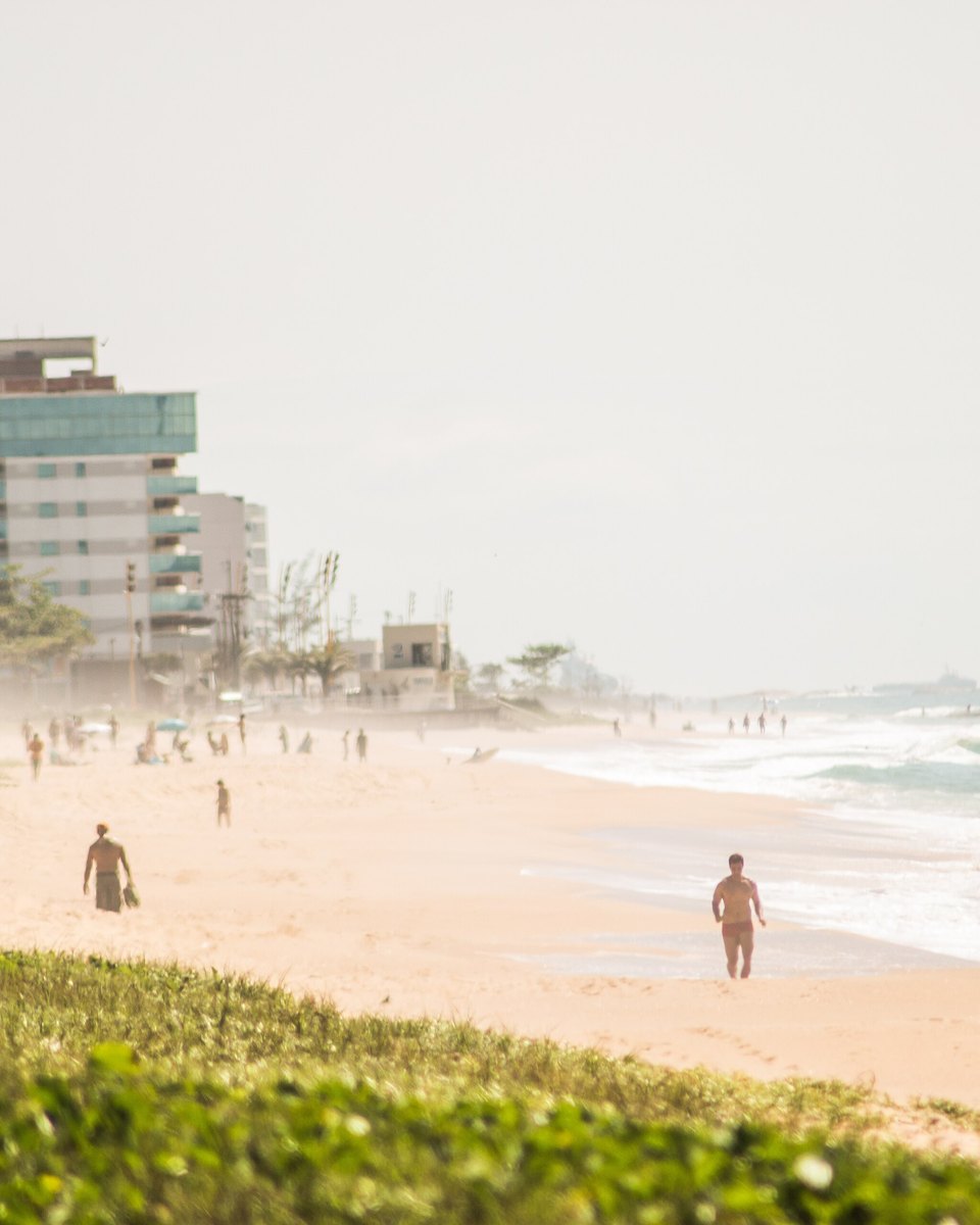 Pessoas caminhando na areia da Praia do Pecado, em Macaé, com prédios à beira-mar.