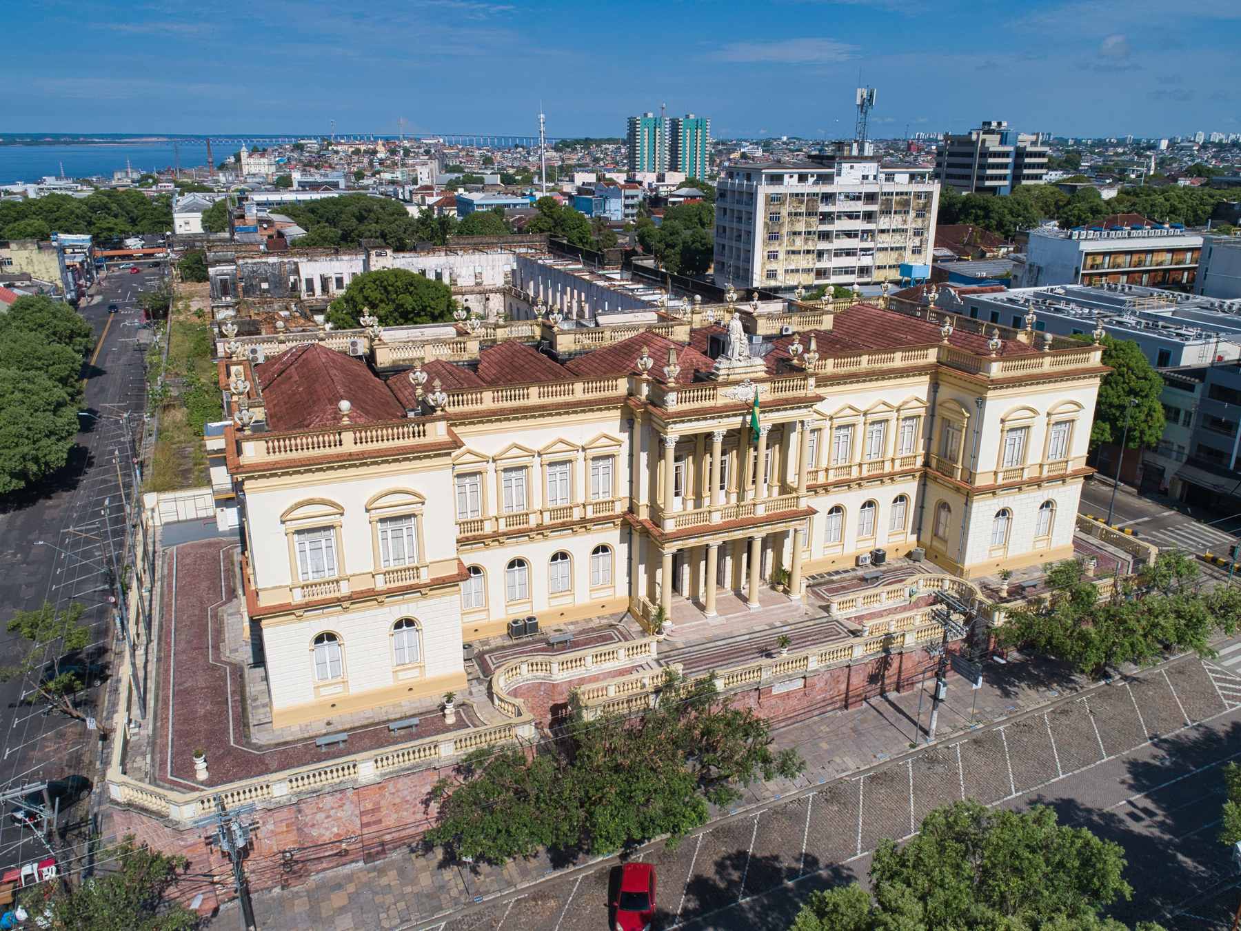 Vista aérea do Palácio da Justiça do Amazonas, prédio neoclássico amarelo e branco no centro de Manaus, com o casario em volta e o rio Negro ao fundo em dia de céu azul