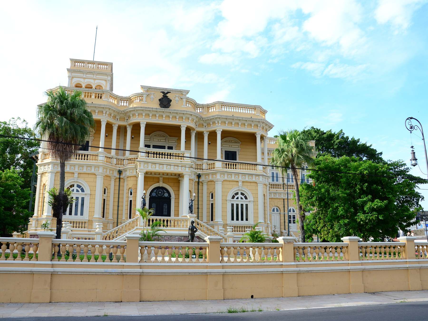 Fachada amarela do Palácio Rio Negro em Manaus, casarão do ciclo da borracha com varanda de colunas brancas e balaustrada, cercado por palmeiras sob céu azul