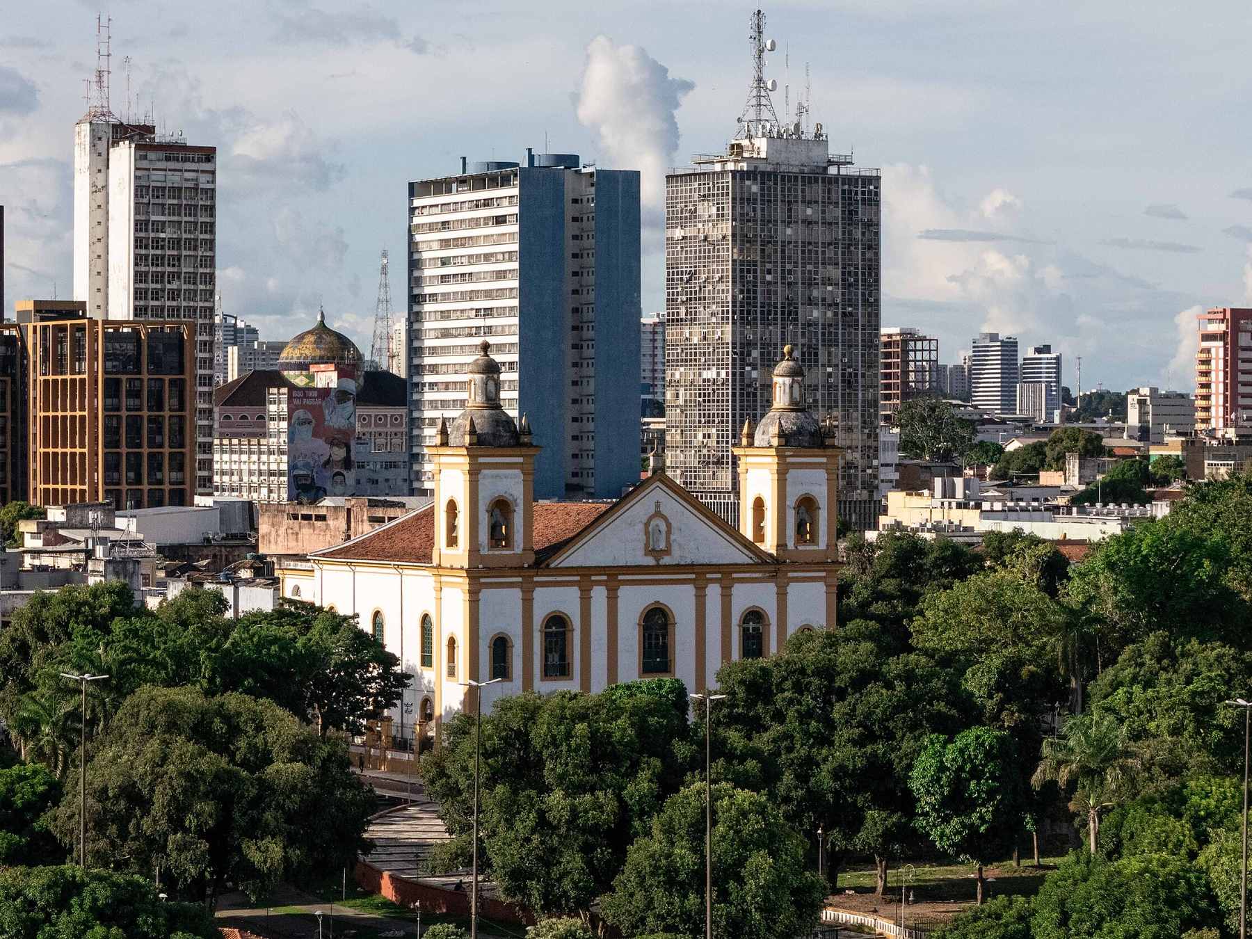 Catedral Metropolitana de Manaus com suas duas torres brancas no meio da praça arborizada, com os prédios do centro da cidade ao fundo