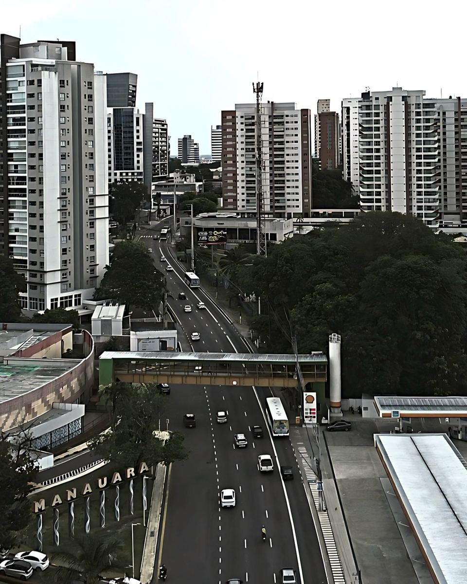 Avenida movimentada no bairro Adrianópolis, em Manaus, com prédios residenciais altos dos dois lados, ônibus, carros e a passarela de pedestres.
