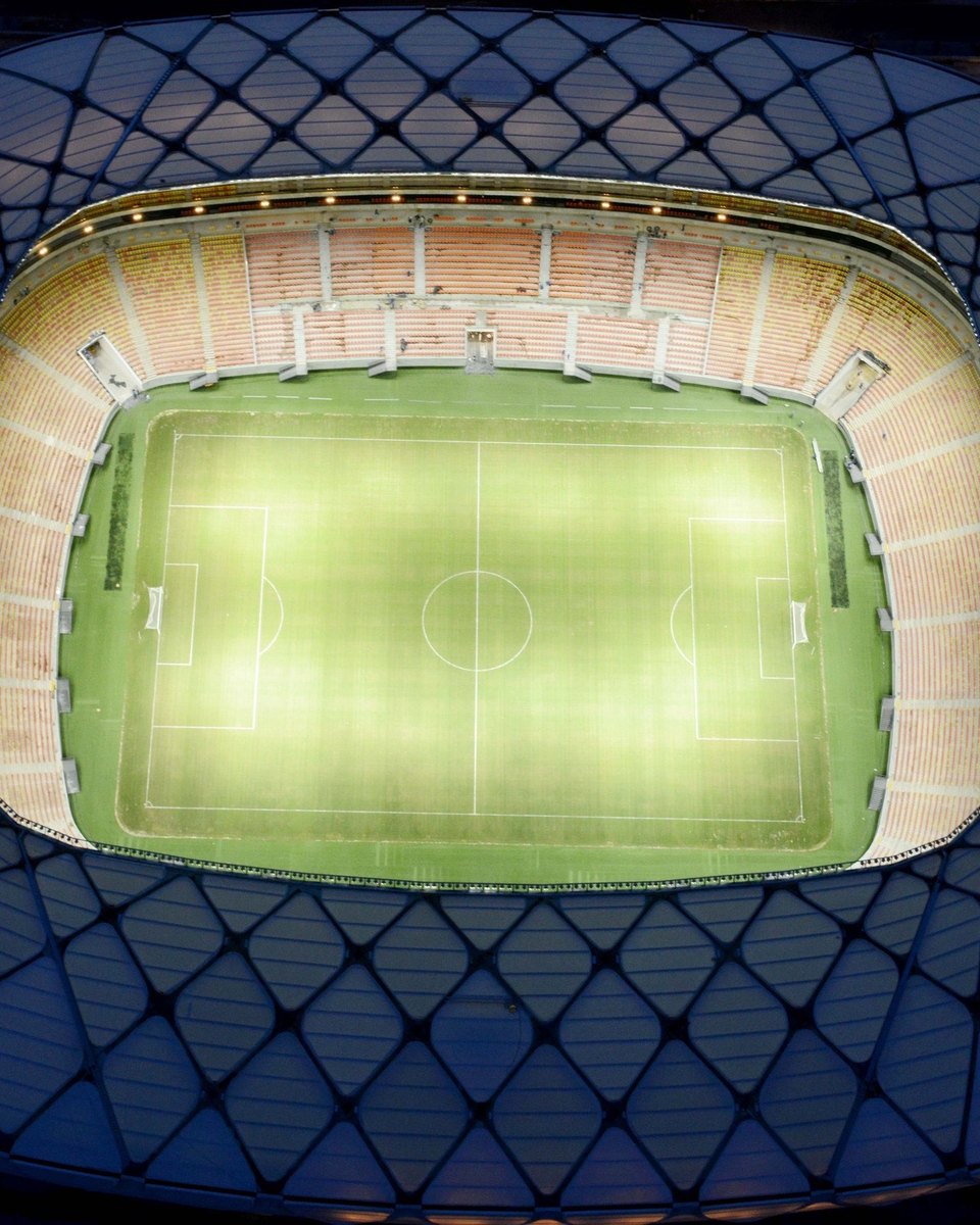 Arena da Amazônia iluminada à noite, vista de cima, com a fachada de treliça metálica e o gramado aceso no centro.
