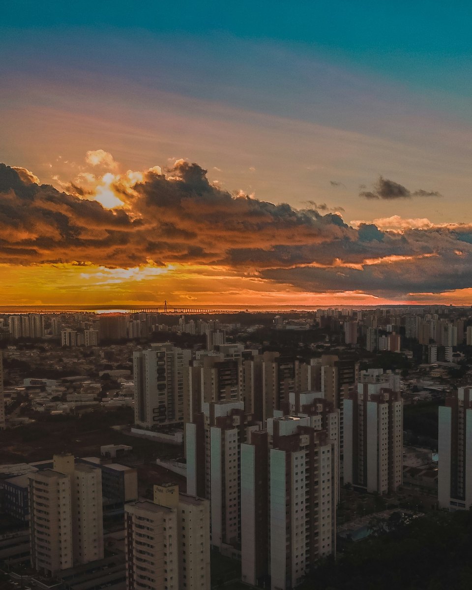 Vista aérea de Manaus no fim da tarde, com os prédios altos da zona centro-sul em primeiro plano e o céu alaranjado sobre o rio Negro.
