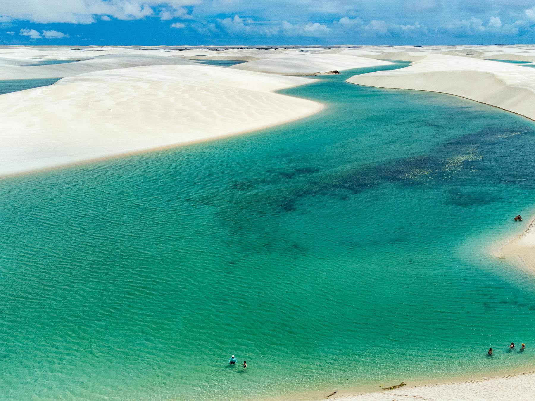 Lagoa de água verde e transparente entre dunas de areia branca no Parque Nacional dos Lençóis Maranhenses, vista do alto, com banhistas na beira da água e o céu azul com nuvens ao fundo