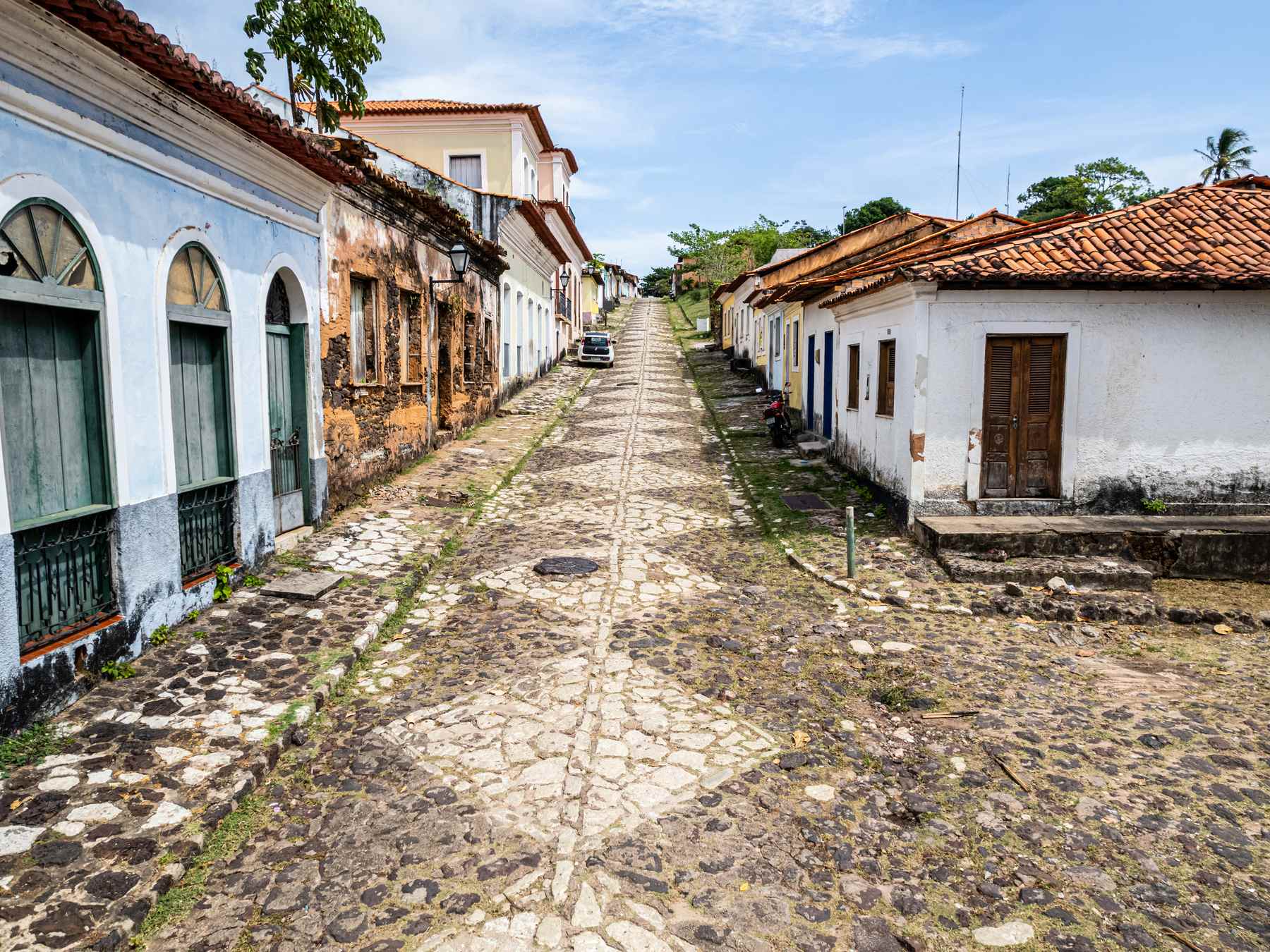 Rua de pedra do centro histórico de Alcântara descendo entre casarões coloniais de telhado de barro, com um sobrado azul e branco à esquerda, casas caiadas à direita e palmeiras ao fundo