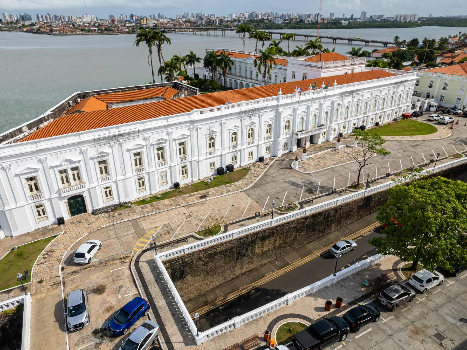 Palácio dos Leões visto do alto em São Luís, com a fachada branca de dois andares e o telhado de barro, o pátio murado com carros estacionados e a Baía de São Marcos com a ponte ao fundo