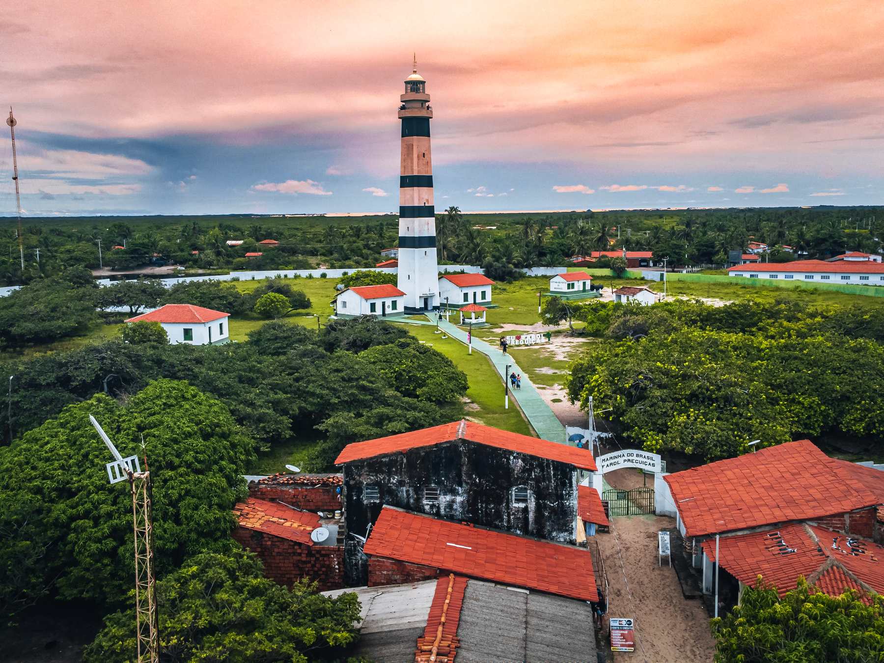 Farol de Mandacaru visto do alto no fim da tarde, com a torre listrada de preto e branco acima do povoado de telhados vermelhos, a vegetação verde em volta e o céu rosado