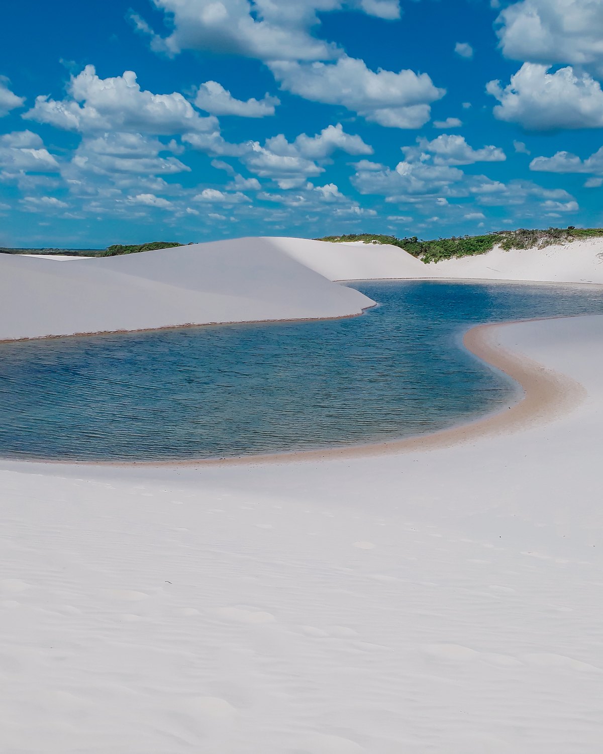 Lagoa de água azul esverdeada cheia entre dunas de areia branca nos Lençóis Maranhenses, com céu de nuvens