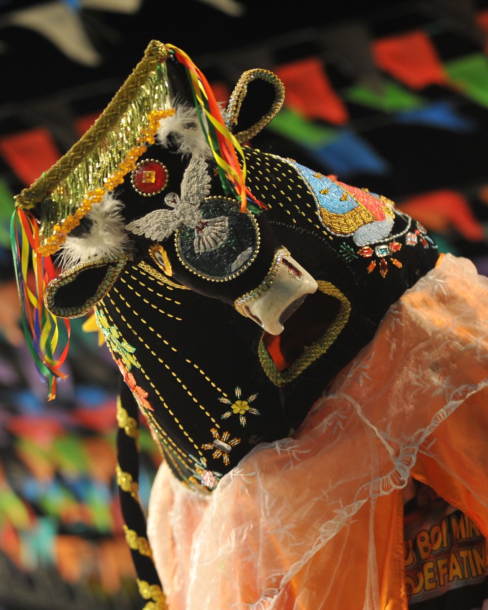 Boi bordado de veludo preto com miçangas, fitas coloridas e capa laranja, erguido por um brincante durante apresentação de bumba meu boi nas festas juninas de São Luís, com bandeirinhas ao fundo