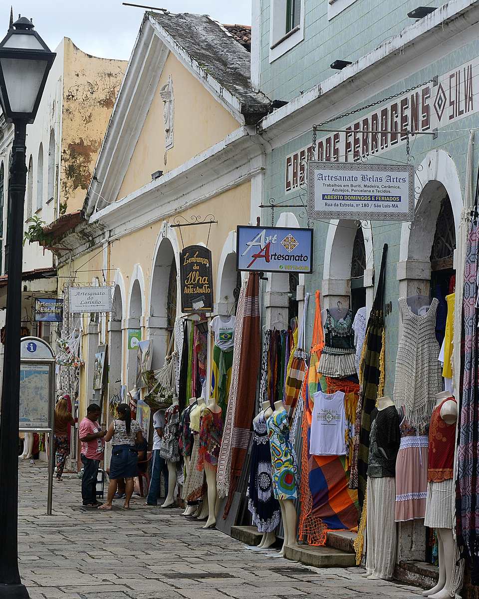 Rua de pedra do bairro da Praia Grande, em São Luís, com lojas de artesanato sob arcadas, redes e roupas penduradas na calçada, placas de comércio e pessoas caminhando