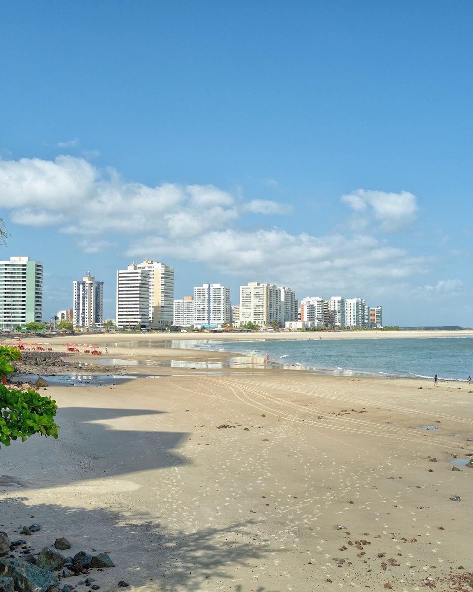 Faixa larga de areia da praia da Ponta d'Areia, em São Luís, com barracas e guarda-sóis na beira, pessoas caminhando perto da água e os prédios do bairro alinhados ao fundo