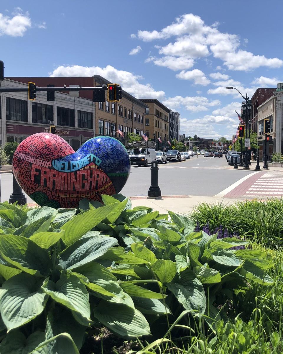 Escultura em forma de coração com a frase Welcome to Framingham, no centro de Framingham, com a faixa de pedestres e os prédios comerciais ao fundo.