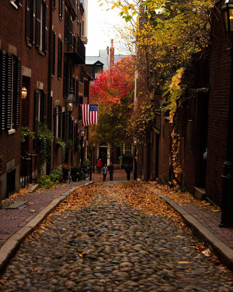 Pessoas caminhando pela Acorn Street, rua de pedras de Beacon Hill, em Boston, coberta de folhas secas no outono, entre casas de tijolo e árvores amarelas e vermelhas.