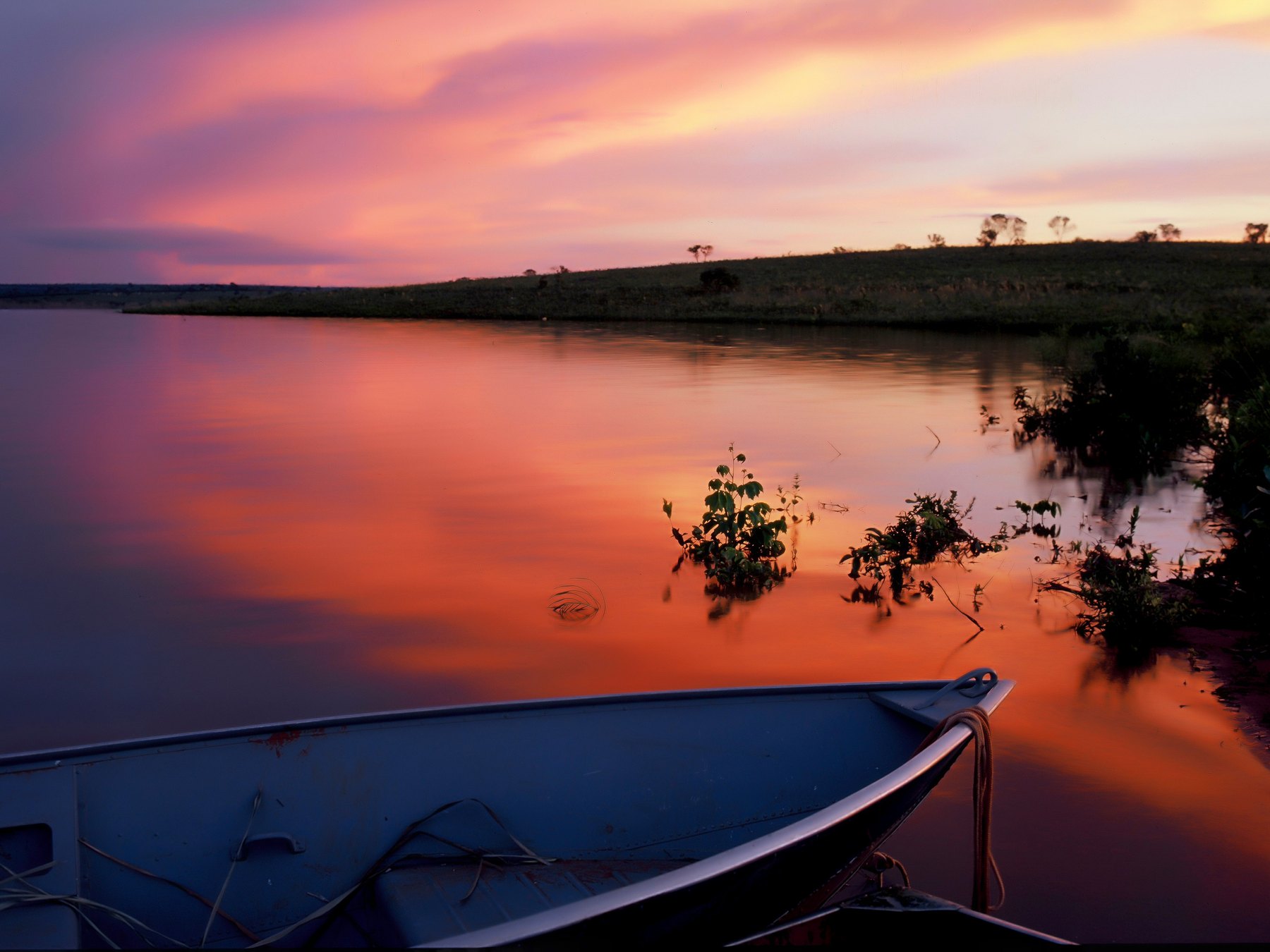 Pôr do sol rosa e alaranjado refletido na água parada do Pantanal, com um barco de alumínio em primeiro plano