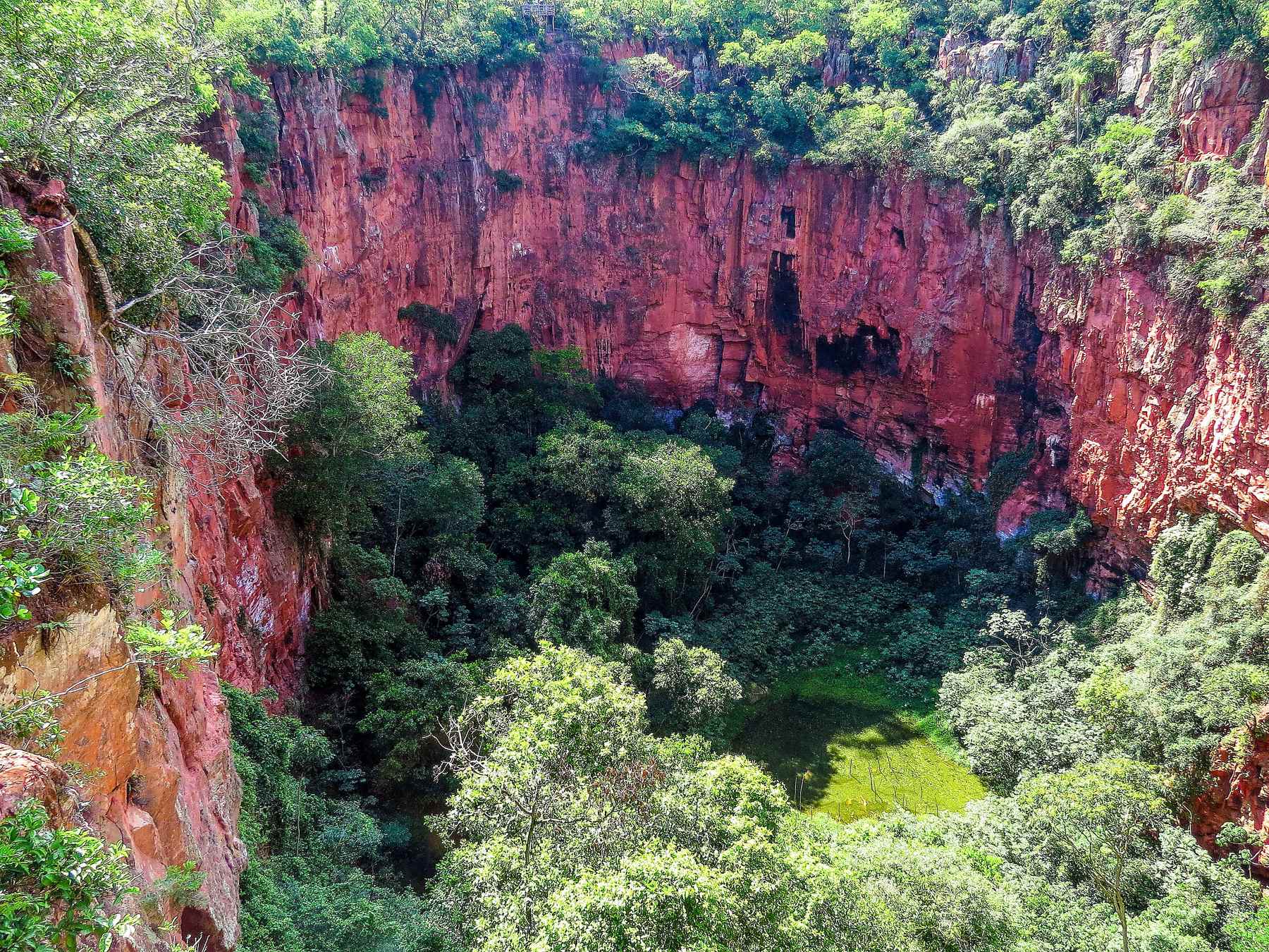 Paredões de rocha vermelha do Buraco das Araras cercando a mata fechada e a lagoa esverdeada no fundo da dolina