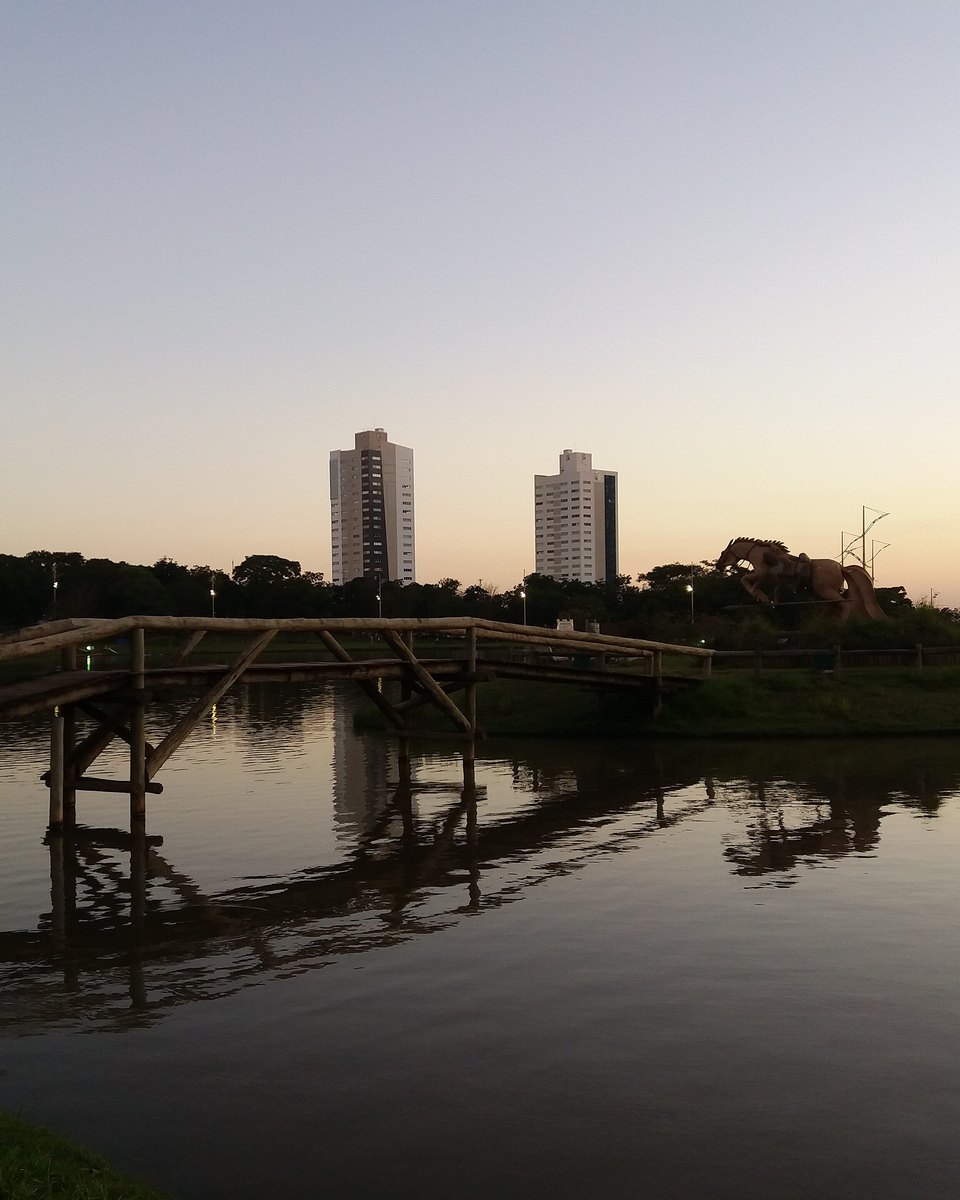 Lago do Parque das Nações Indígenas, em Campo Grande, com a ponte de madeira atravessando a água e os prédios da cidade recortados no céu do fim de tarde.
