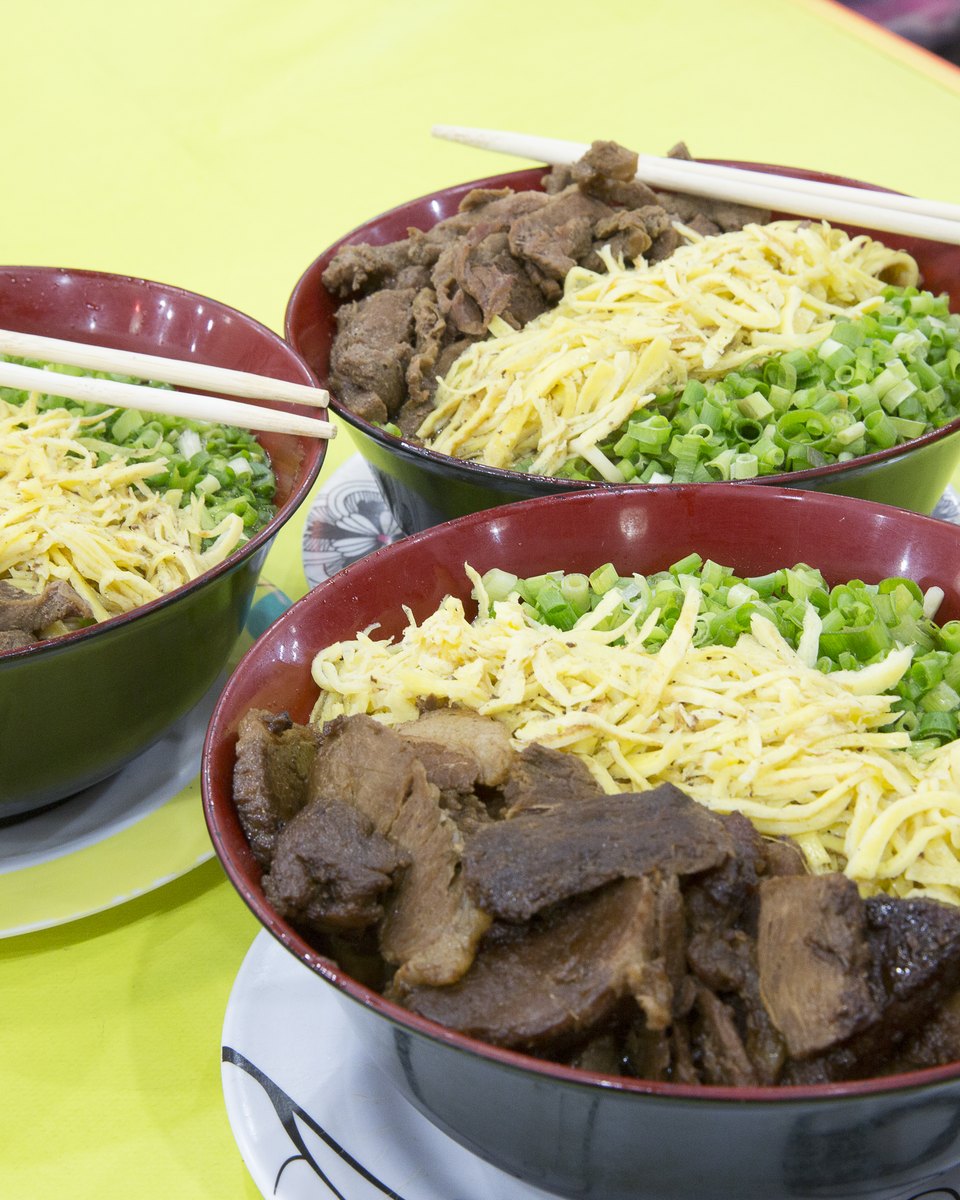 Três tigelas de sobá com macarrão, ovo em tiras, carne e cebolinha sobre uma mesa amarela, com hashis apoiados nas bordas.