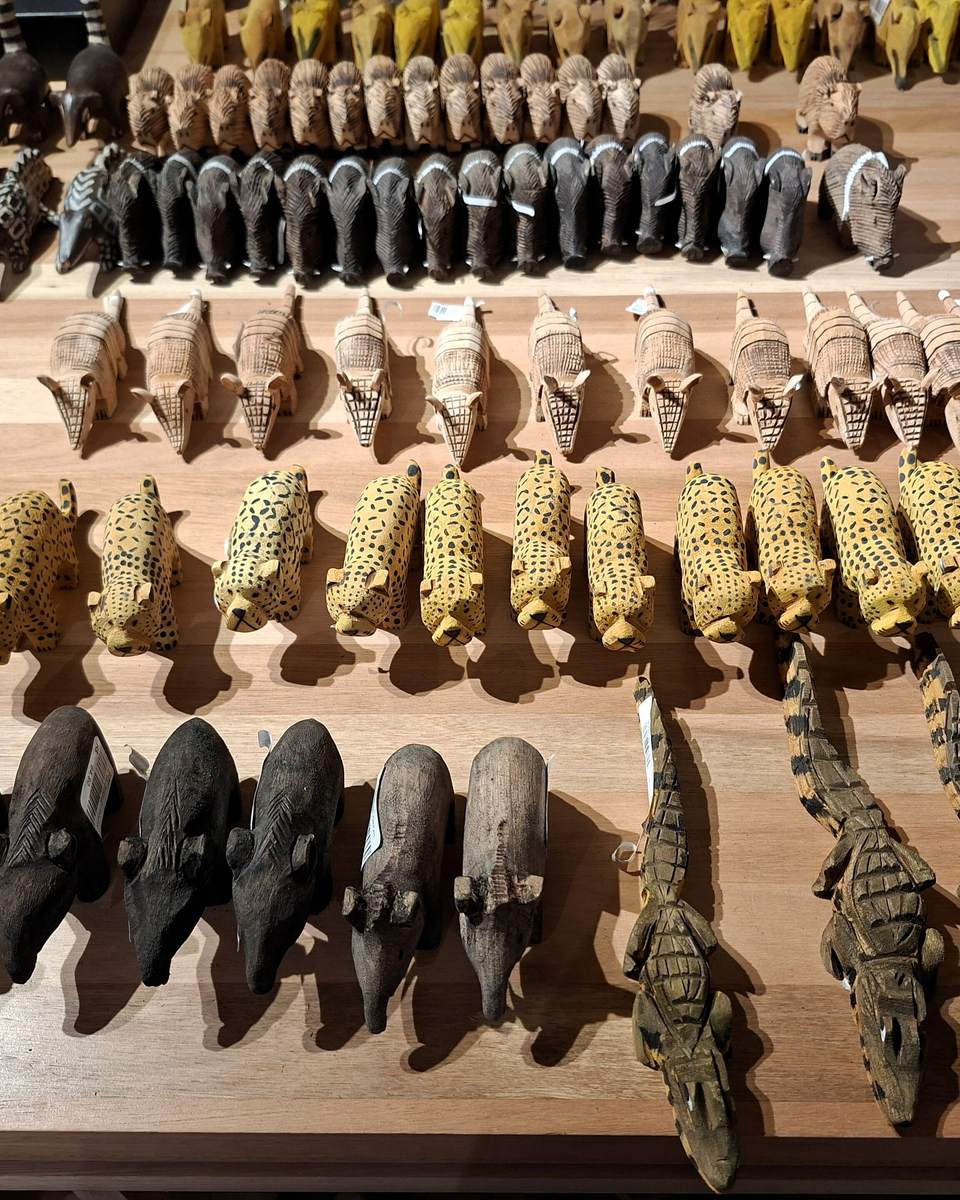 Fileiras de animais do Pantanal esculpidos em madeira e pintados à mão, artesanato do povo Terena, expostos em uma mesa com etiquetas de preço.