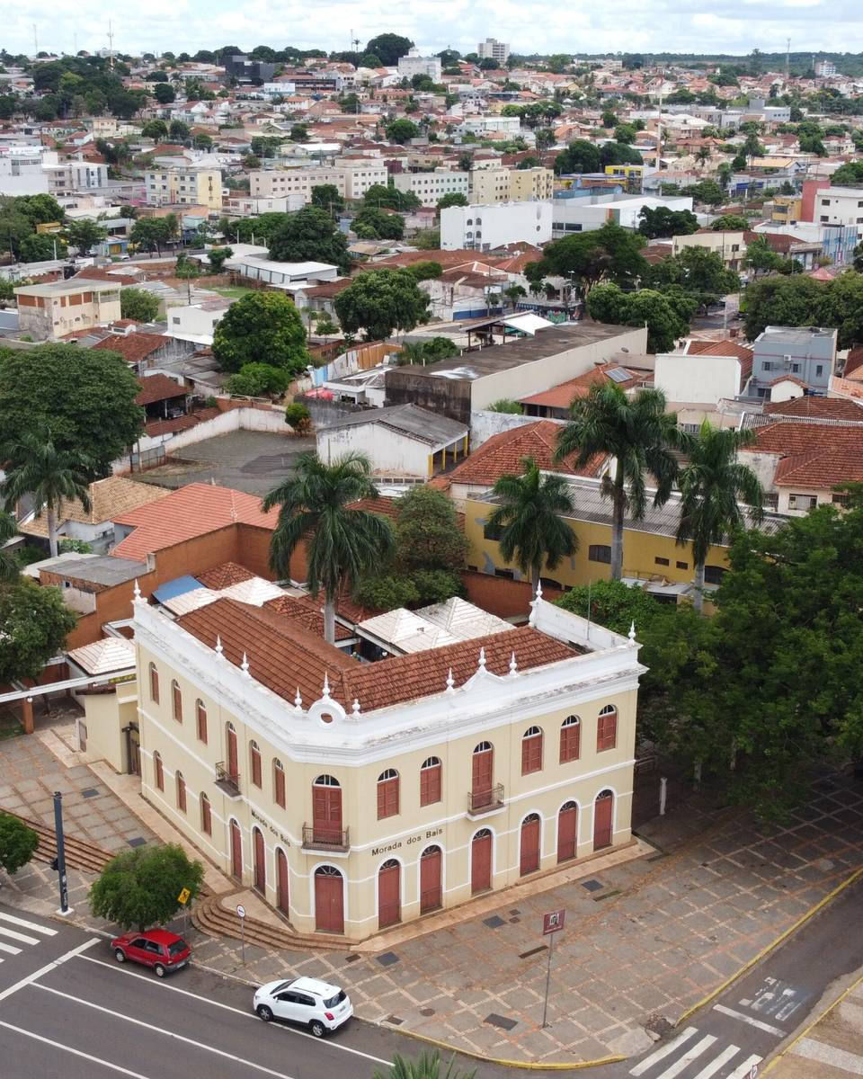 Vista aérea da Morada dos Baís, o casarão amarelo do centro de Campo Grande, com a avenida em frente e os telhados da cidade se espalhando até o horizonte.