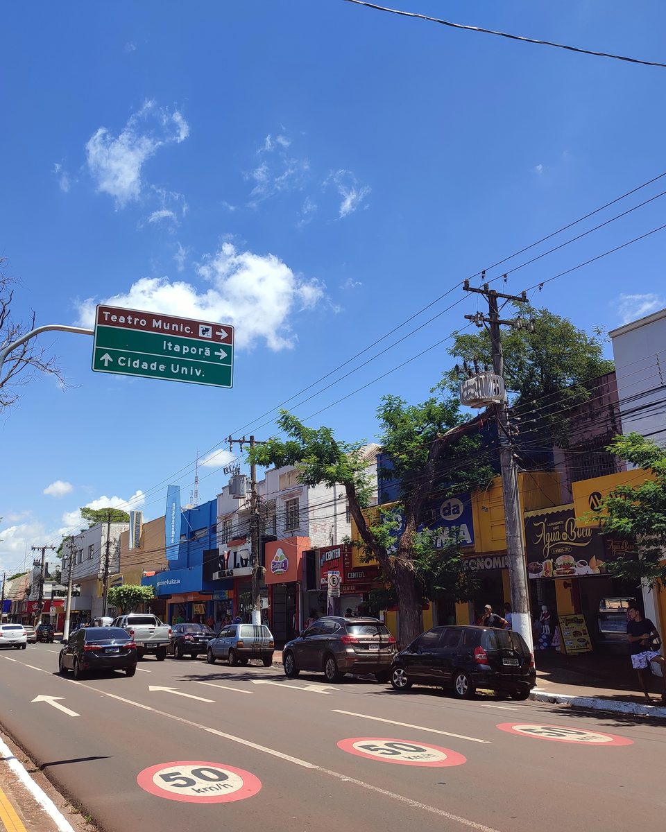 Avenida Marcelino Pires no centro de Dourados, com lojas, carros no trânsito e placa indicando o Teatro Municipal.