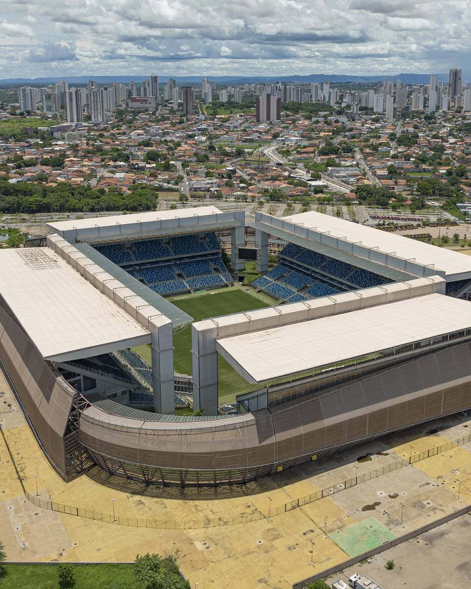 Vista aérea da Arena Pantanal, em Cuiabá, com a cobertura branca do estádio, os prédios da cidade em volta e o cerrado no horizonte.
