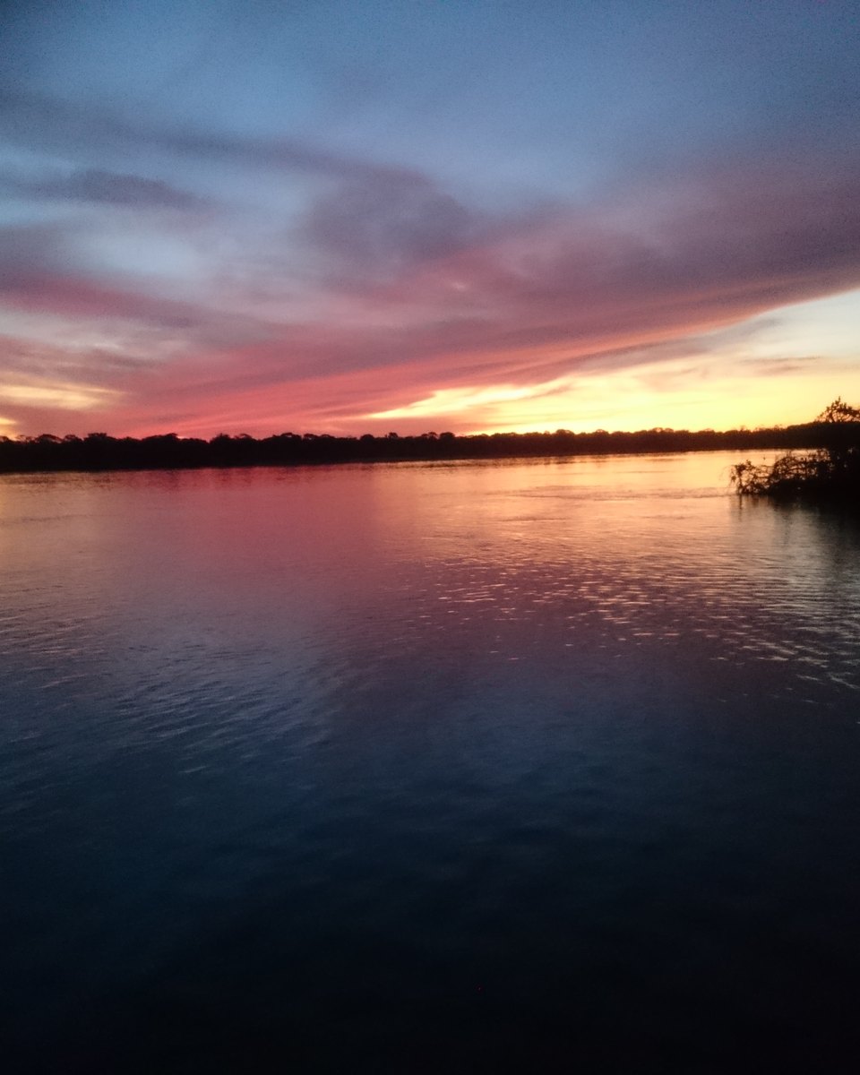 Entardecer na beira do rio Xingu, dentro do Parque Indígena do Xingu, com a mata em silhueta nas duas margens e o céu alaranjado refletido na água.