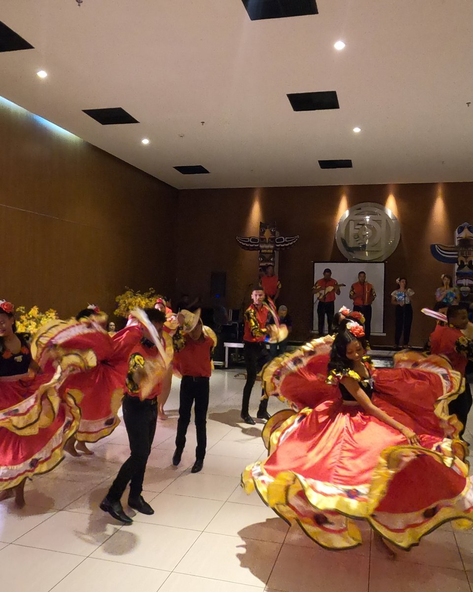 Dançarinas de saia rodada vermelha e amarela e dançarinos de chapéu se apresentando em uma roda de siriri em Cuiabá, com os músicos tocando ao fundo.