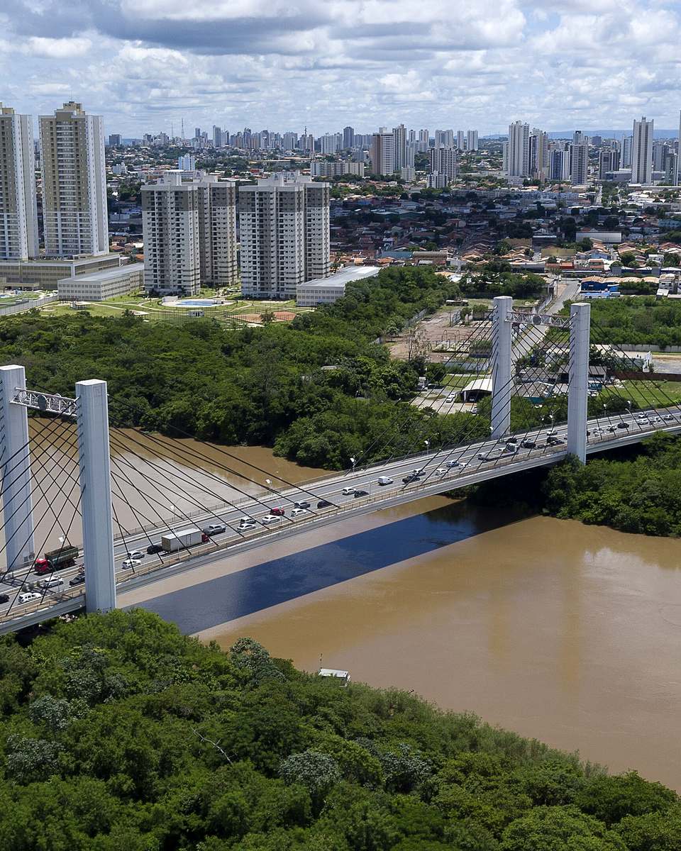 Vista aérea da ponte Sérgio Motta cruzando o rio Cuiabá entre Várzea Grande e Cuiabá, com mata nas margens e os prédios das duas cidades ao fundo.