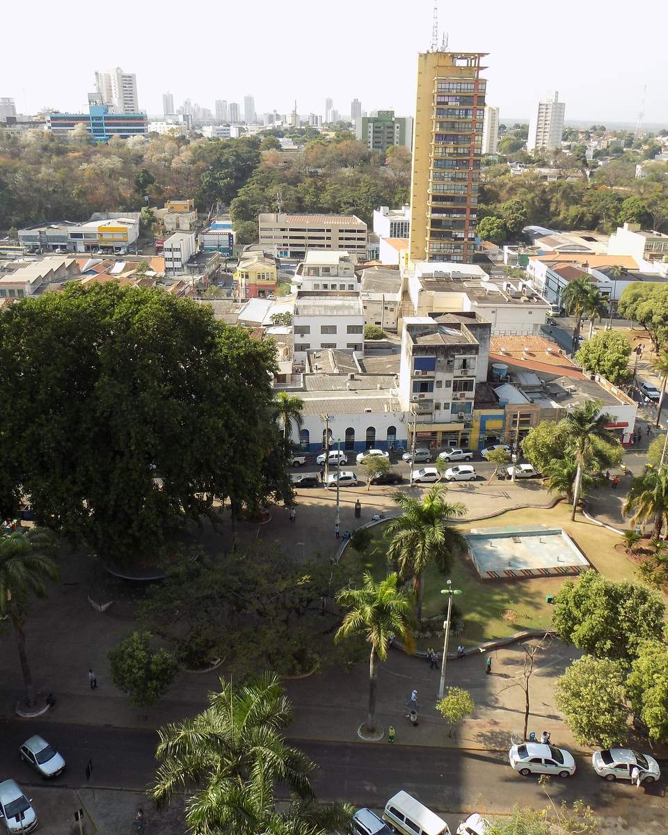 Centro de Cuiabá visto do alto em tarde de sol forte, com palmeiras, as árvores da praça e carros estacionados ao longo da rua.