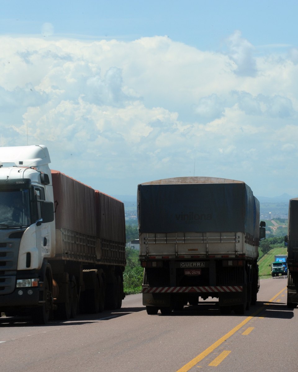 Caminhões graneleiros carregados seguindo em fila pela rodovia BR-163 na saída de Rondonópolis, com o relevo da cidade ao fundo.