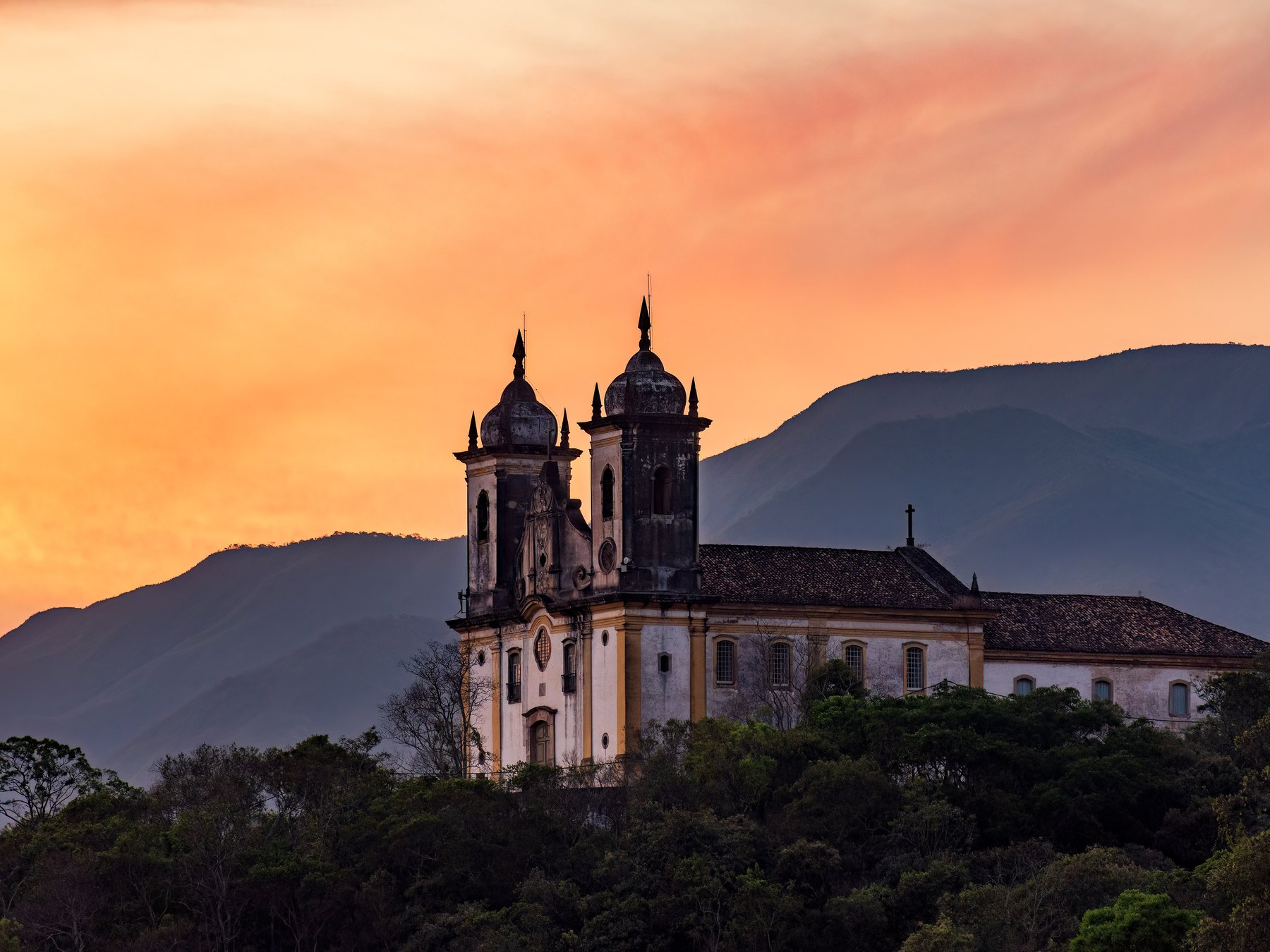 Igreja de São Francisco de Paula no alto do morro em Ouro Preto, com as montanhas ao fundo e o céu laranja do entardecer.