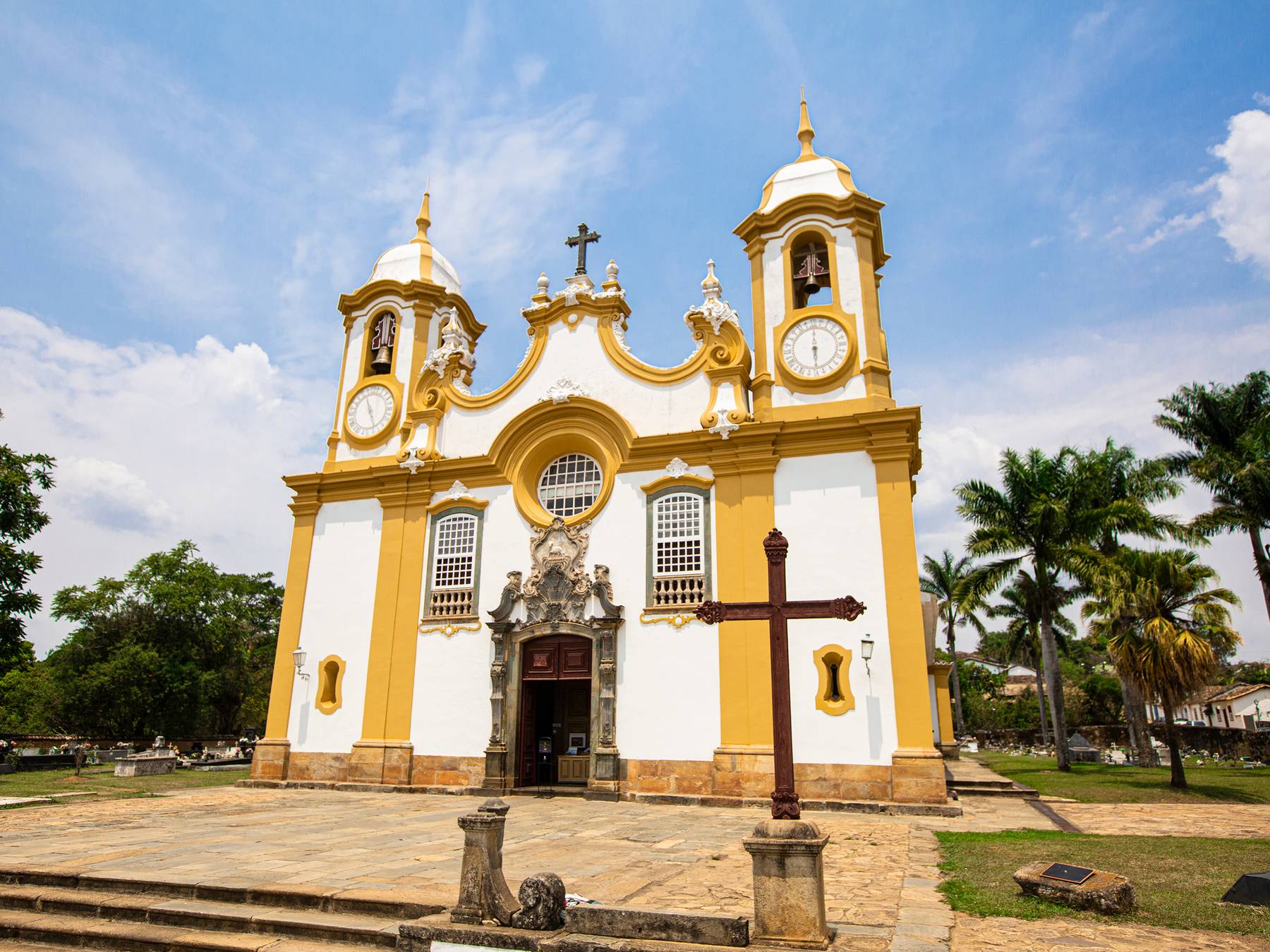 Fachada branca e amarela da Matriz de Santo Antônio, com palmeiras e céu azul, em Tiradentes.