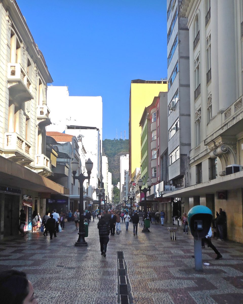 Pedestres caminhando pelo calçadão da Rua Halfeld, no centro de Juiz de Fora, entre prédios antigos e lojas.