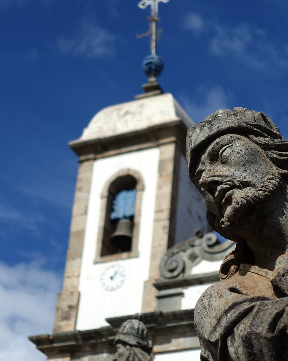 Profeta de pedra-sabão esculpido por Aleijadinho em primeiro plano, com as torres brancas da Basílica do Bom Jesus de Matosinhos contra o céu azul, em Congonhas.