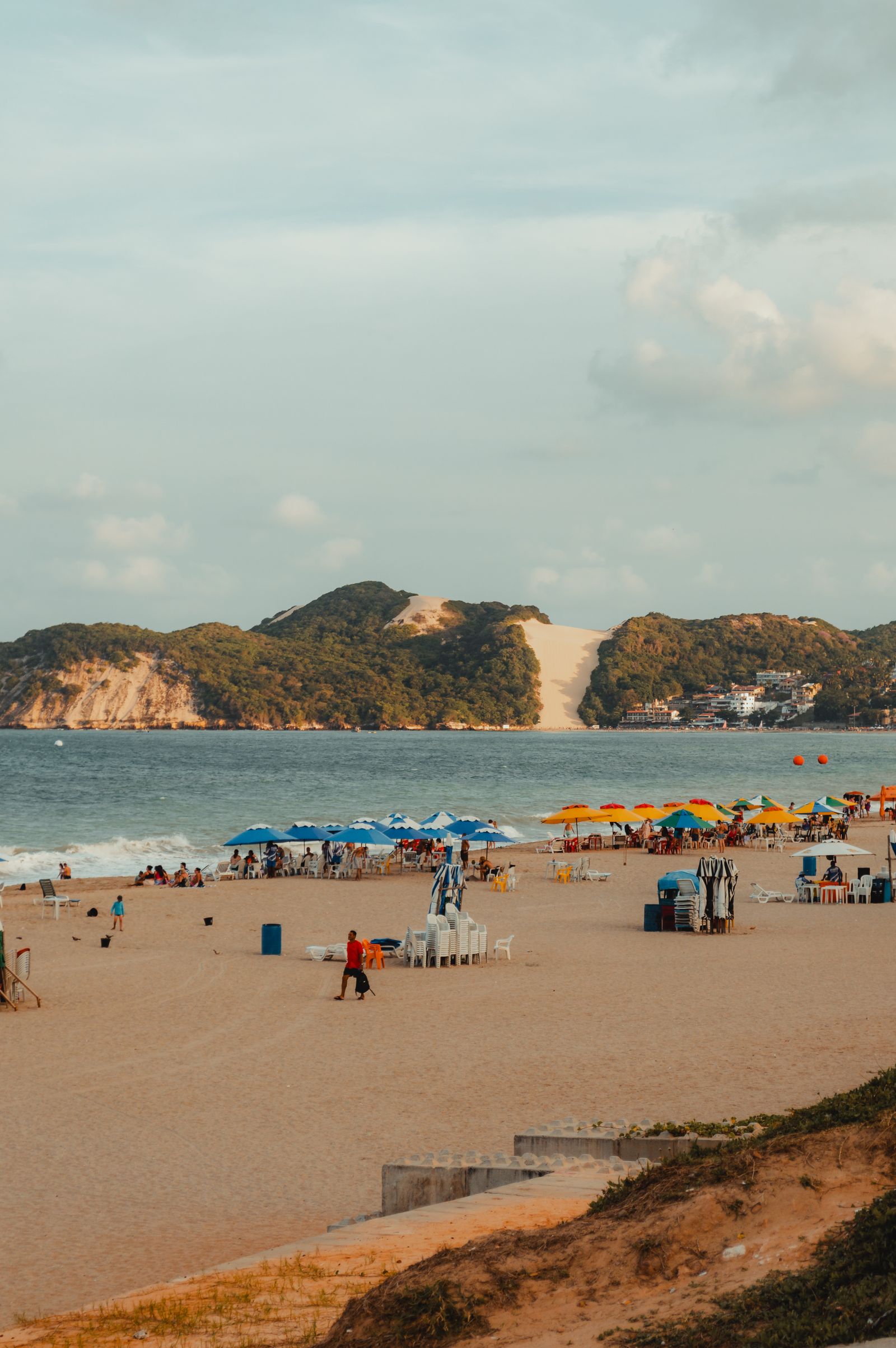 Praia de Ponta Negra cheia de guarda-sóis coloridos e cadeiras de praia, com o Morro do Careca ao fundo entre a vegetação