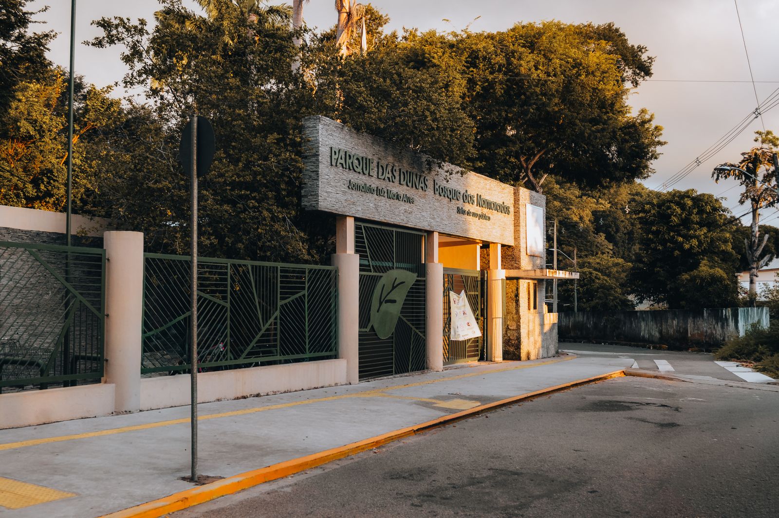 Entrada do Parque das Dunas, Bosque dos Namorados, com placa de identificação entre árvores altas ao entardecer