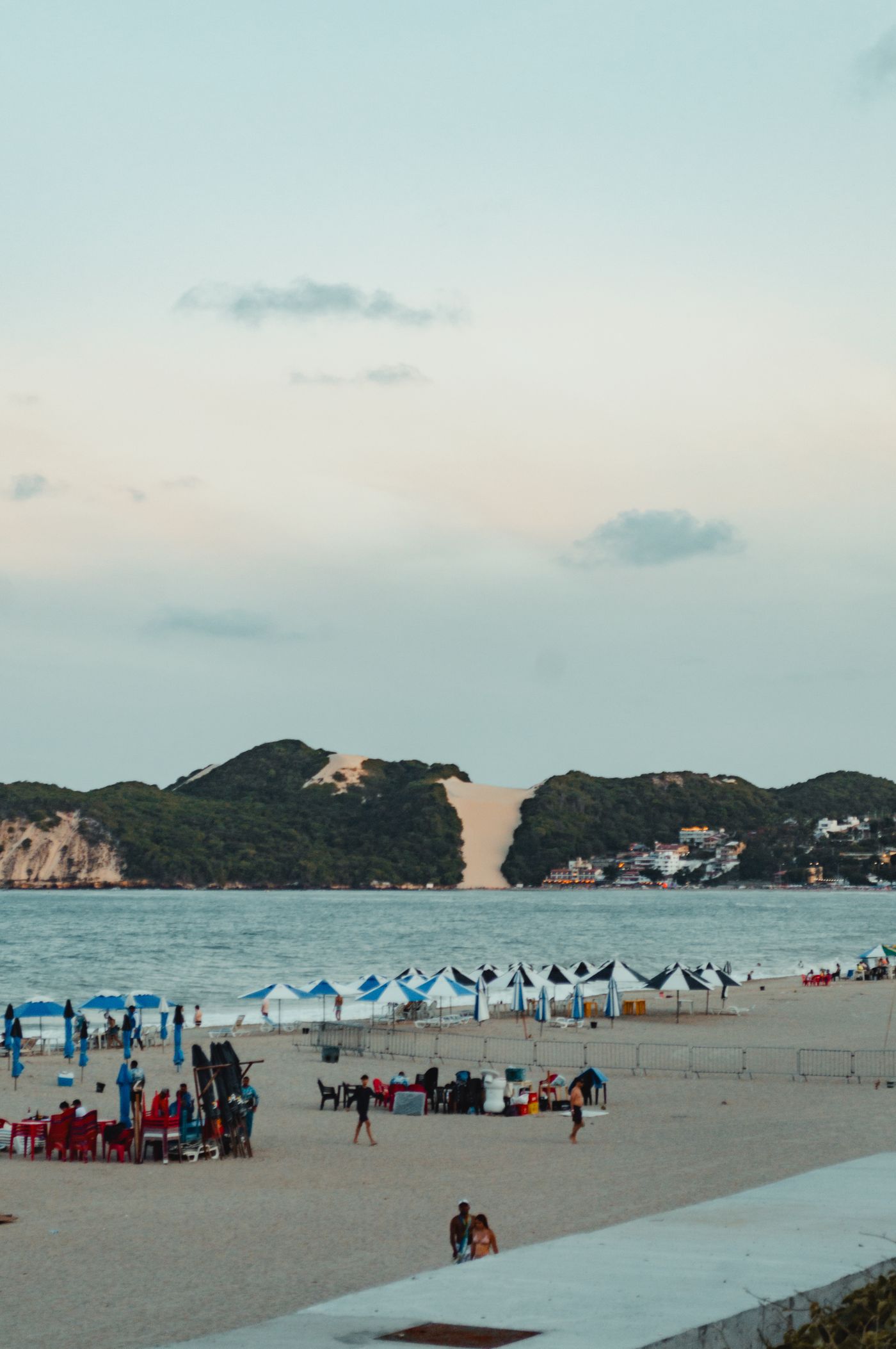 Praia de Ponta Negra ao entardecer, com guarda-sóis na areia e o Morro do Careca, a duna clara entre a vegetação, ao fundo