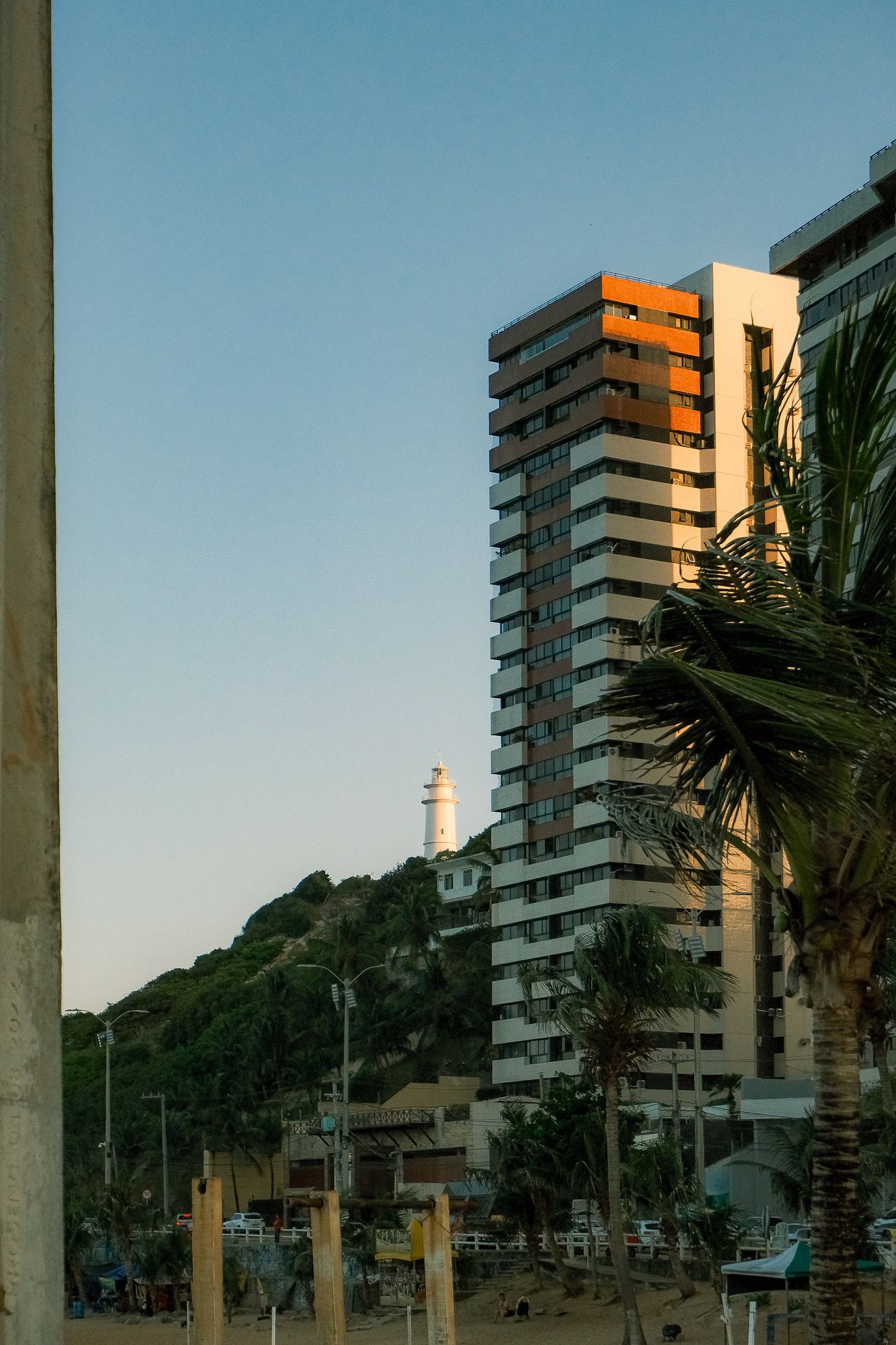 Farol de Mãe Luíza visto ao longe, no alto de uma duna coberta de vegetação, com prédios em primeiro plano em Natal