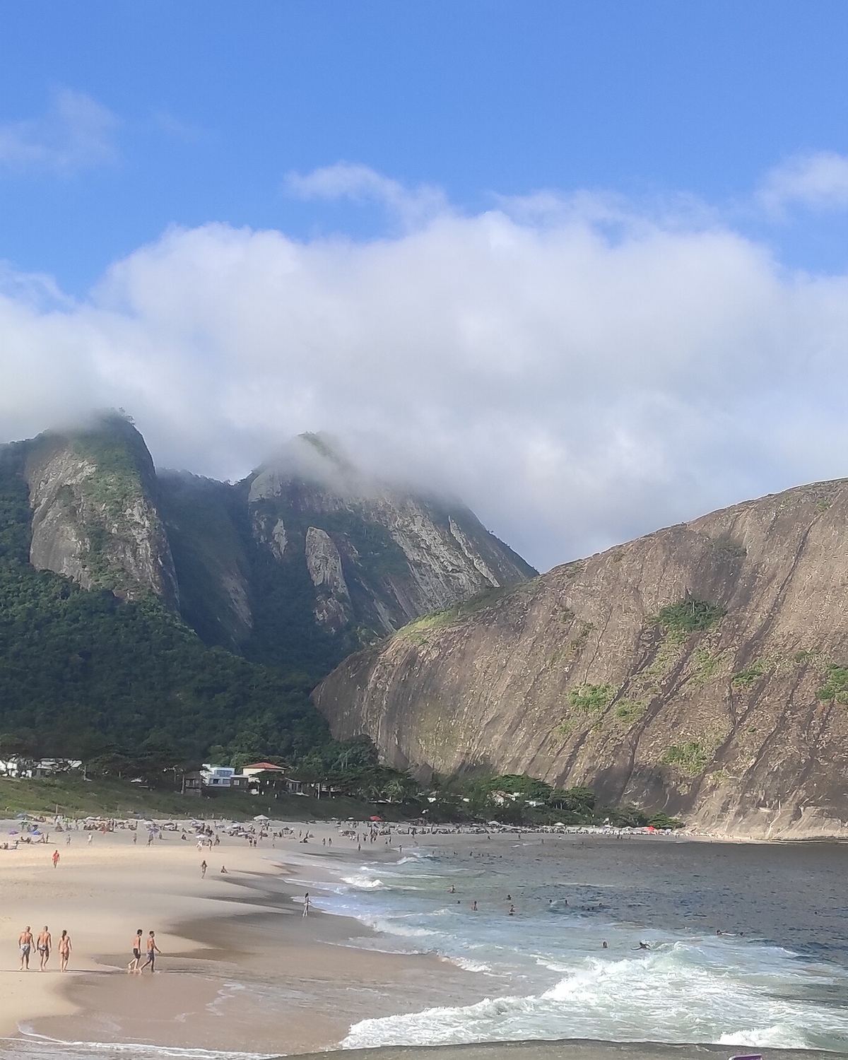 Praia de Itacoatiara, na Região Oceânica de Niterói, com banhistas na areia, ondas quebrando e os costões de pedra cobertos de mata ao fundo, sob nuvens baixas.