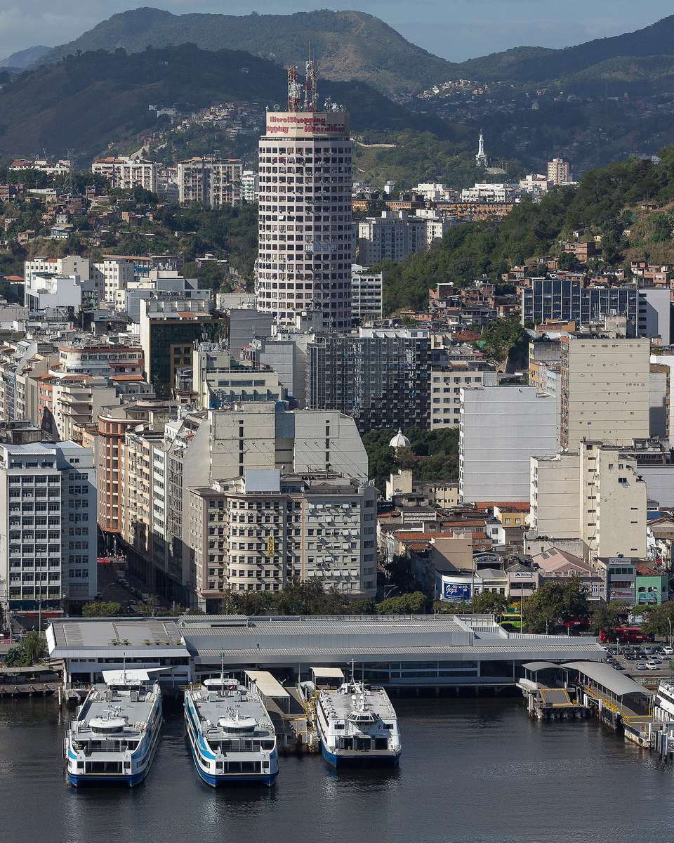 Vista aérea da Estação Praça Arariboia, no Centro de Niterói, com três barcas atracadas na baía, prédios do Centro e os morros da cidade ao fundo.