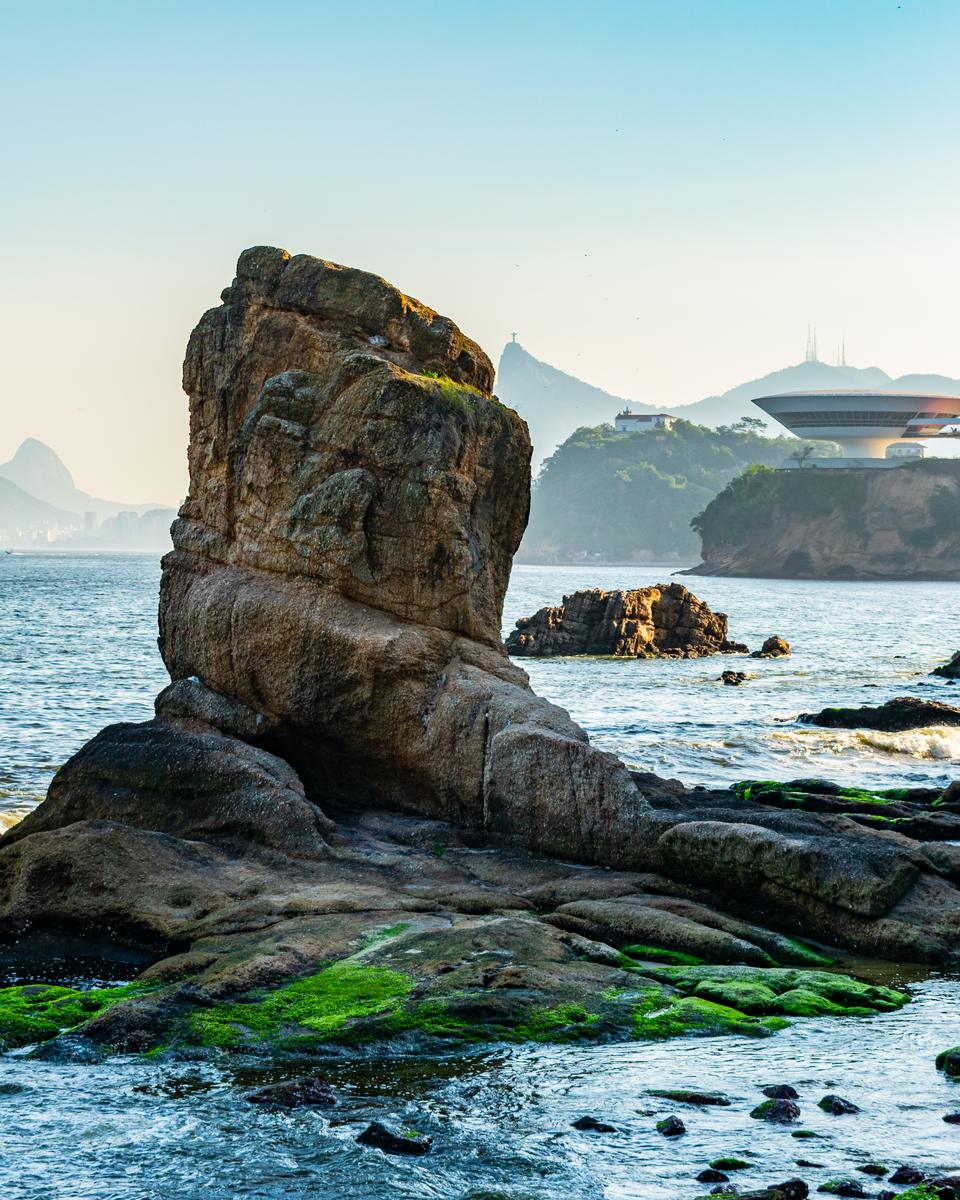 Rocha coberta de limo na orla da Boa Viagem, em Niterói, com o MAC e as montanhas do Rio ao fundo.