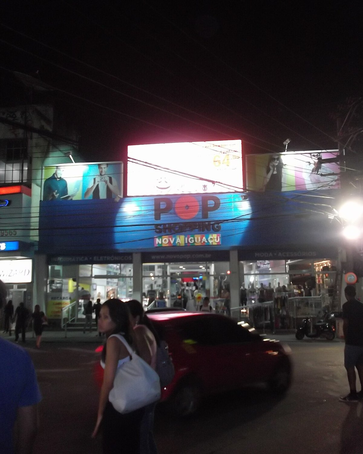 Rua do Centro de Nova Iguaçu à noite, em frente ao TopShopping, com pessoas e lojas iluminadas.