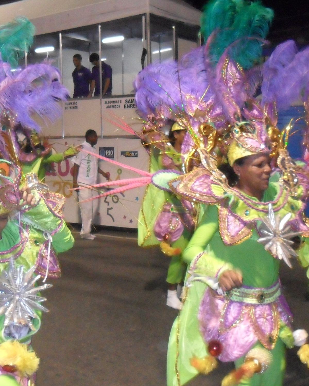 Ala do GRES Império da Uva, escola de samba de Nova Iguaçu, desfilando à noite com fantasias verdes e cocares de penas lilás.