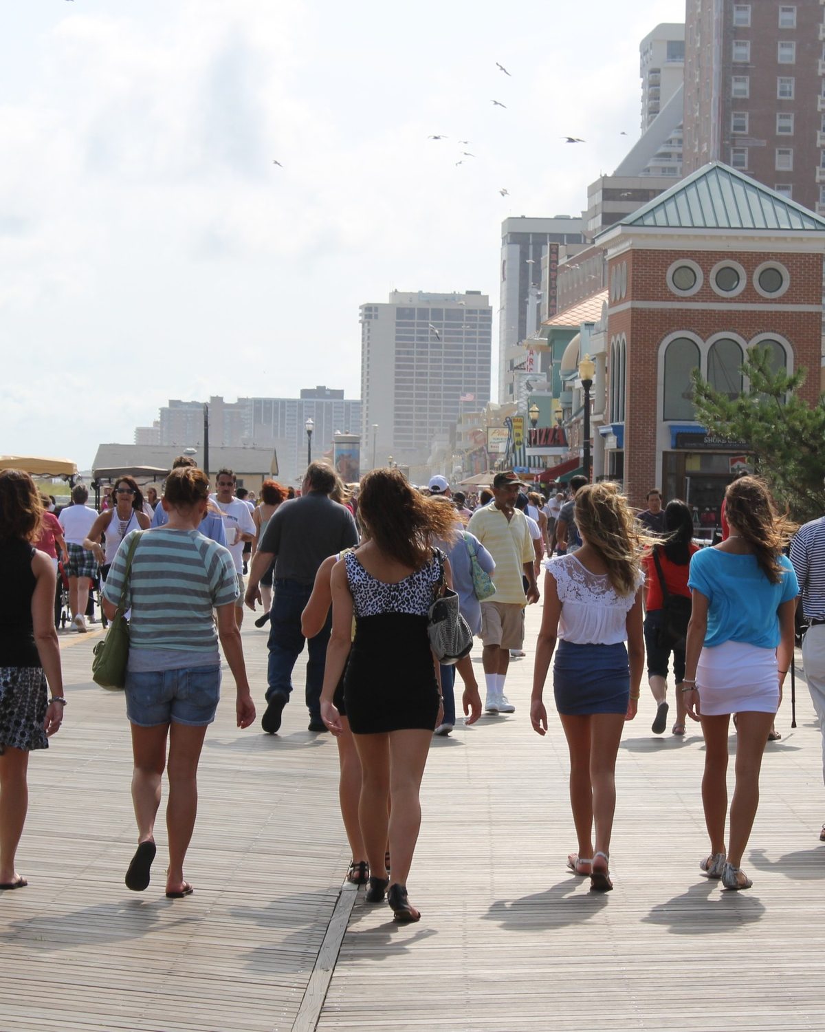 Grupo de pessoas de roupa de verão caminhando de costas pelo calçadão de madeira de Atlantic City, com hotéis e prédios altos ao fundo.