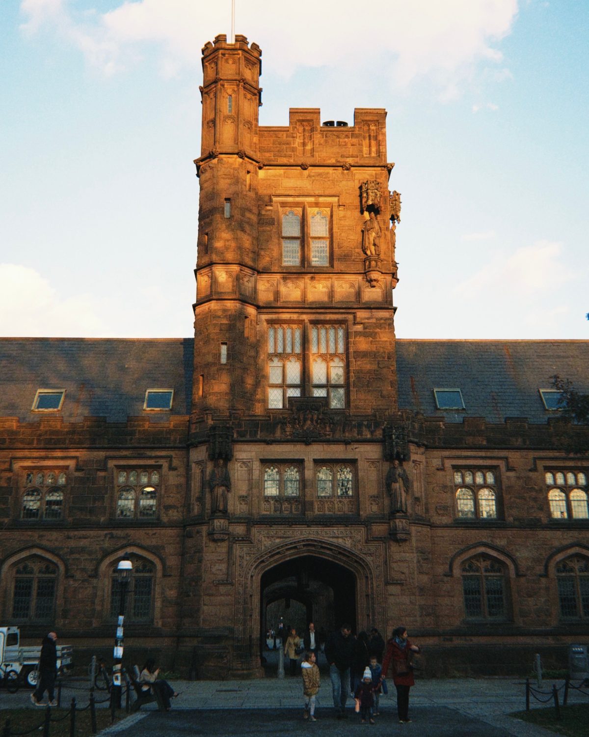 Prédio gótico de pedra com torre na Universidade de Princeton, iluminada pelo sol do fim de tarde, com estudantes passando pela entrada em arco.