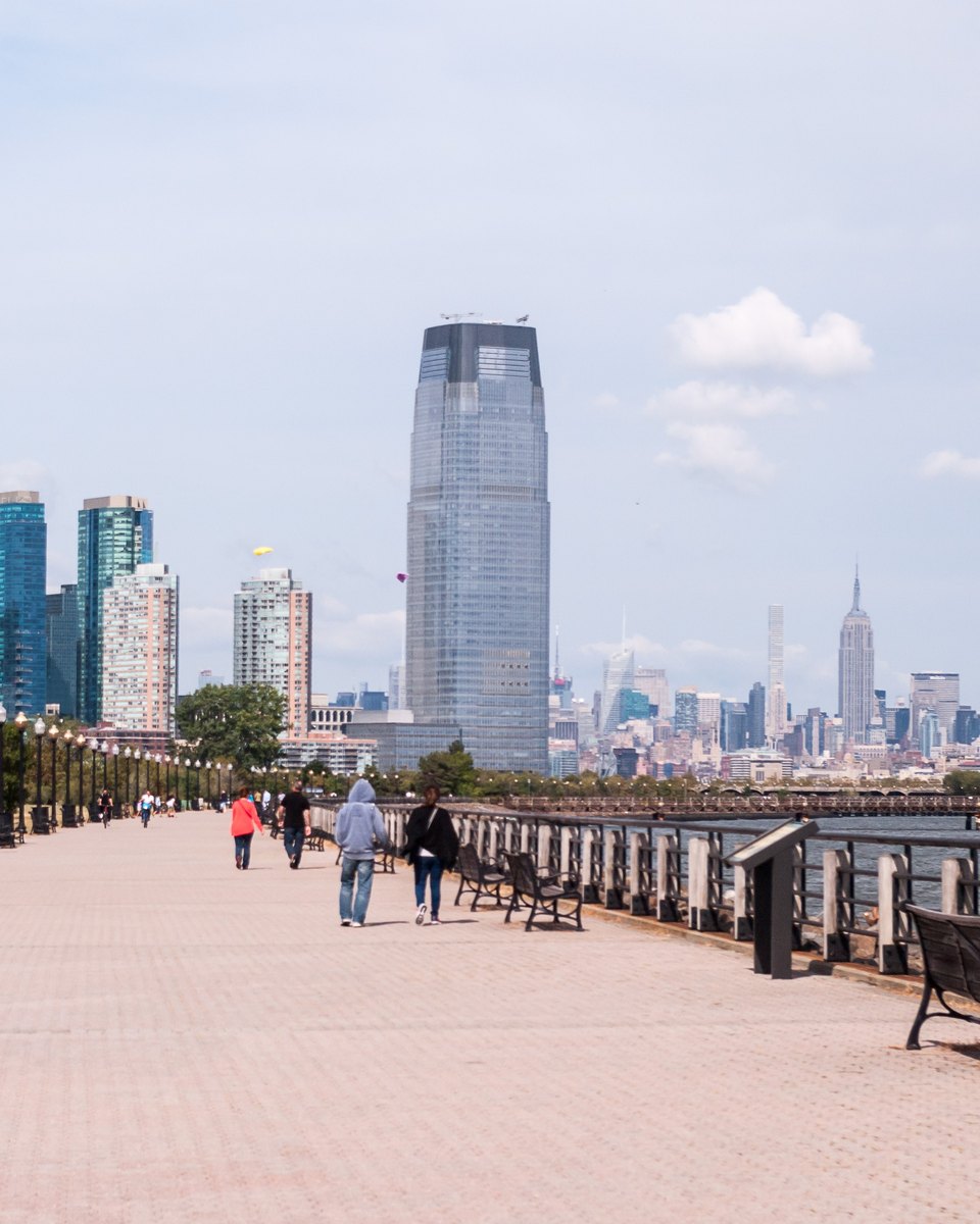 Pessoas caminhando no calçadão à beira do rio no Liberty State Park, em Jersey City, com a torre de vidro da Goldman Sachs e o Empire State Building ao fundo.
